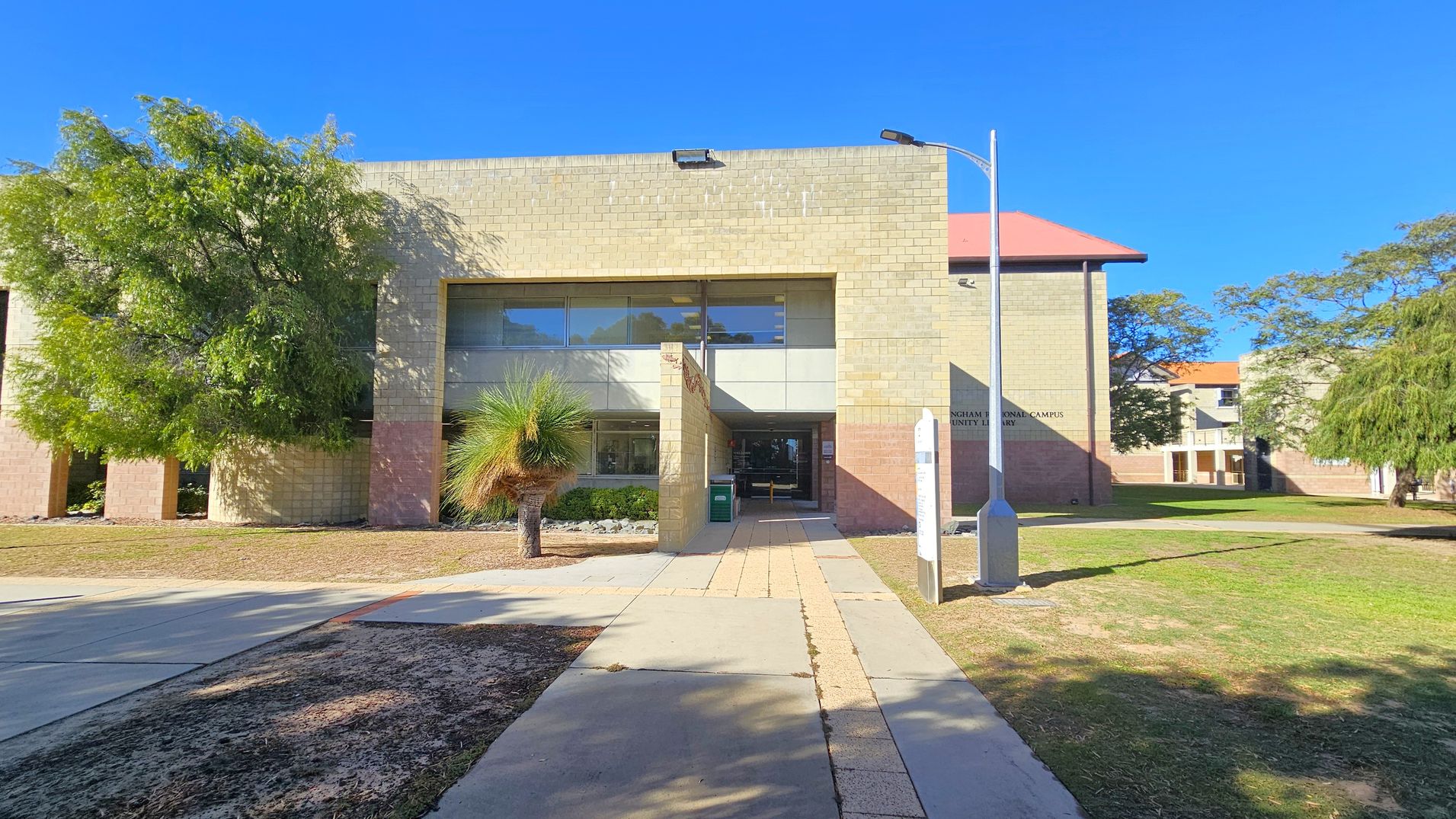 A building with a beige brick facade and large windows in the centre. A pathway leads to the entrance, flanked by a small tree on the left and a lamp post on the right. The building has a flat roof, and there are trees on both sides, with grass and patches of dirt in the foreground. Another building with a red roof is partially visible in the background to the right.
