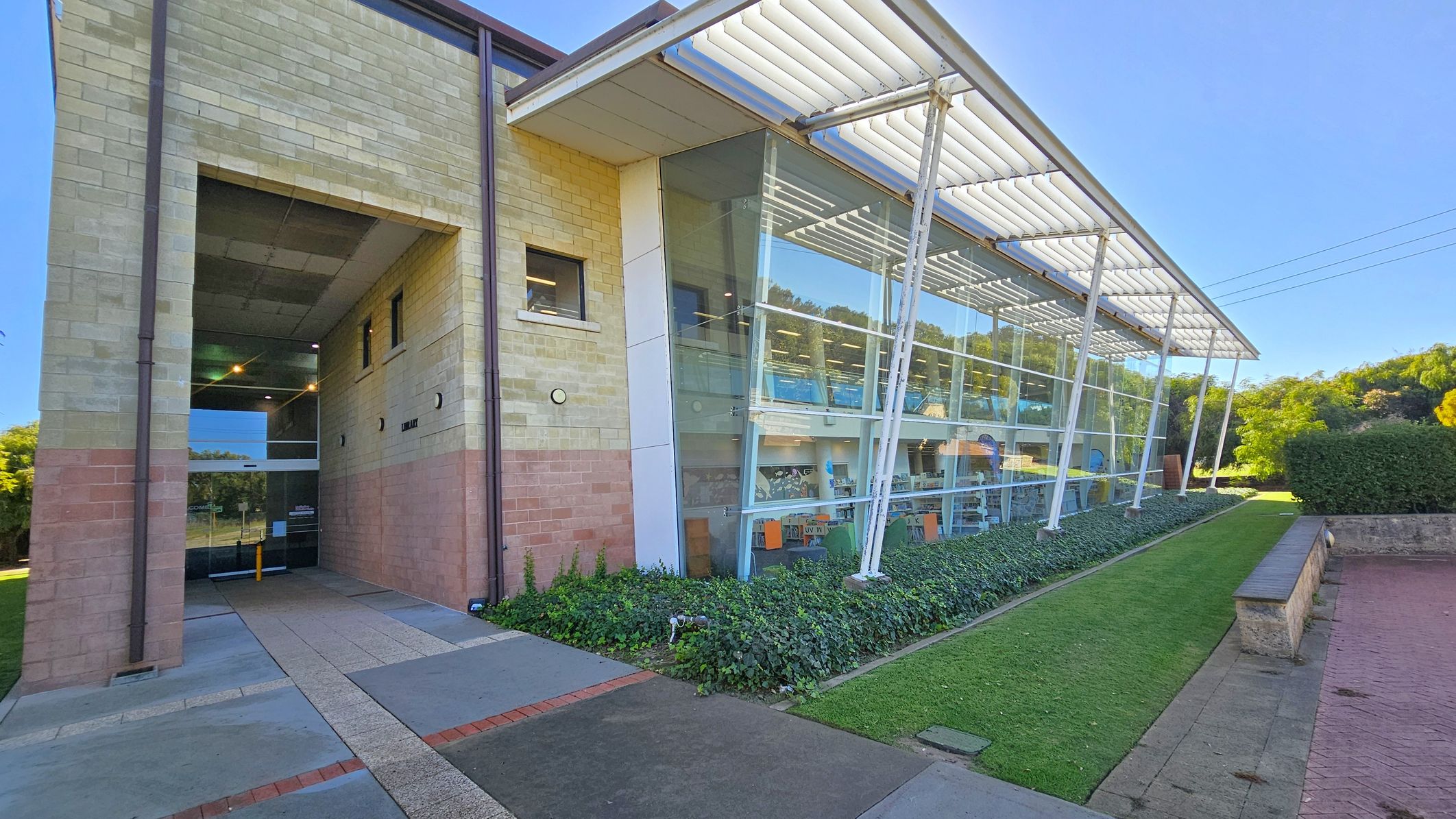 A modern building with a mix of brick and glass. On the left, there's a brick wall with a recessed entrance featuring glass doors. The right side has large glass windows supported by white pillars, allowing a view of the interior. The foreground has a paved path and a small grassy area with low shrubs. A hedge and trees are visible in the background.