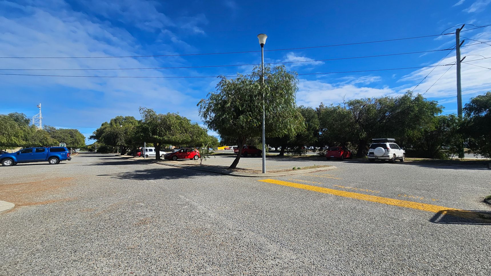 A car park with a gravel surface. On the left, a blue vehicle is parked, while on the right, a white vehicle is visible. In the centre, a tall streetlamp stands next to a tree. Several other trees and parked cars are scattered throughout the background. Power lines stretch across the top of the image, and a yellow painted line is visible on the ground in the foreground.