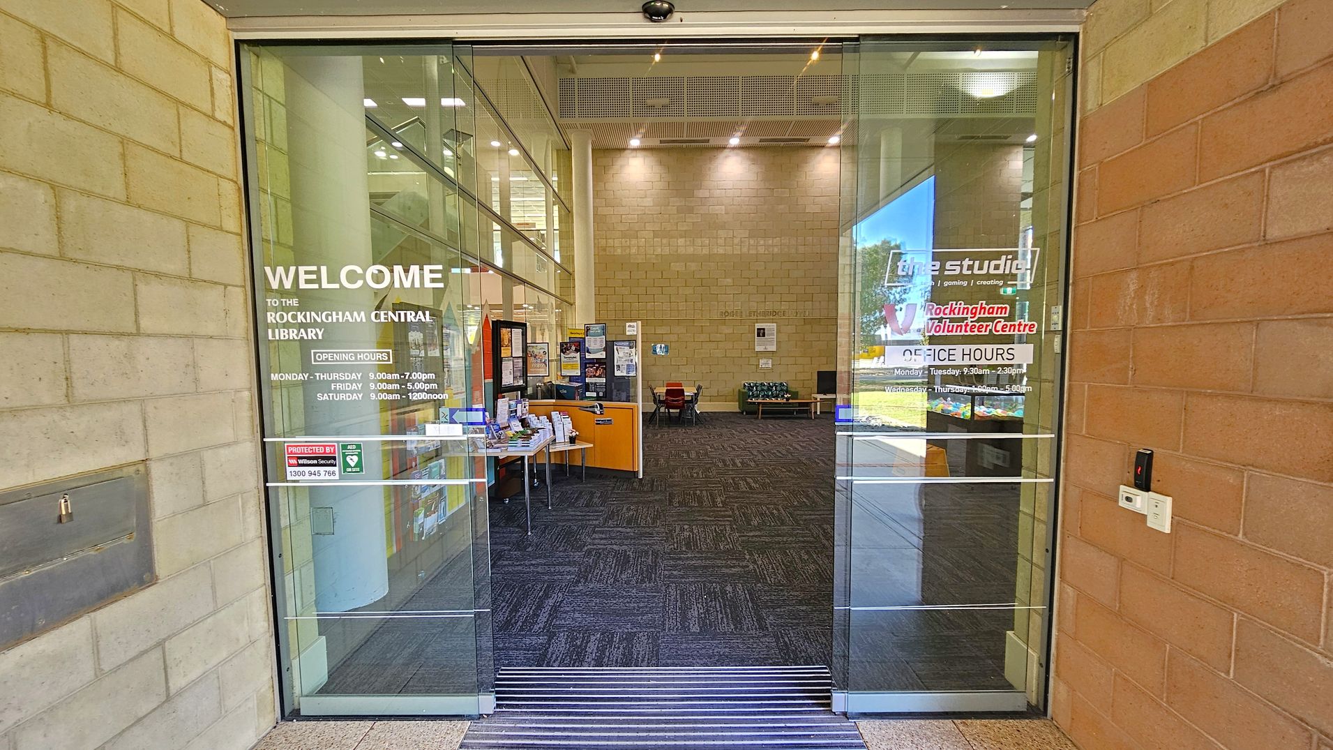 Glass entrance doors to Rockingham Central Library with opening hours displayed. Inside, a spacious area with a dark carpeted floor and light-coloured brick walls. On the left, a small table with brochures and a notice board. In the background, a few chairs and a table are visible. The ceiling is high with recessed lighting. On the right, a window shows a view of the outside.