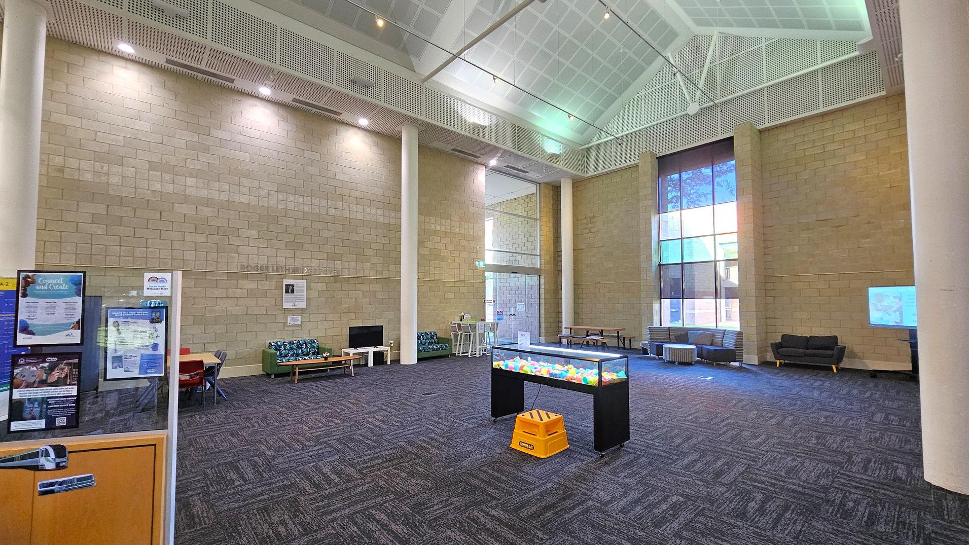 A spacious interior with high ceilings and a grid pattern. The left side features a notice board with posters and a small seating area with a green sofa and a table. In the centre, there's a display table with colourful objects and a yellow step stool. The right side has large windows allowing natural light, with grey sofas and a small table. The floor is carpeted in a dark pattern, and the walls are made of light-coloured bricks.