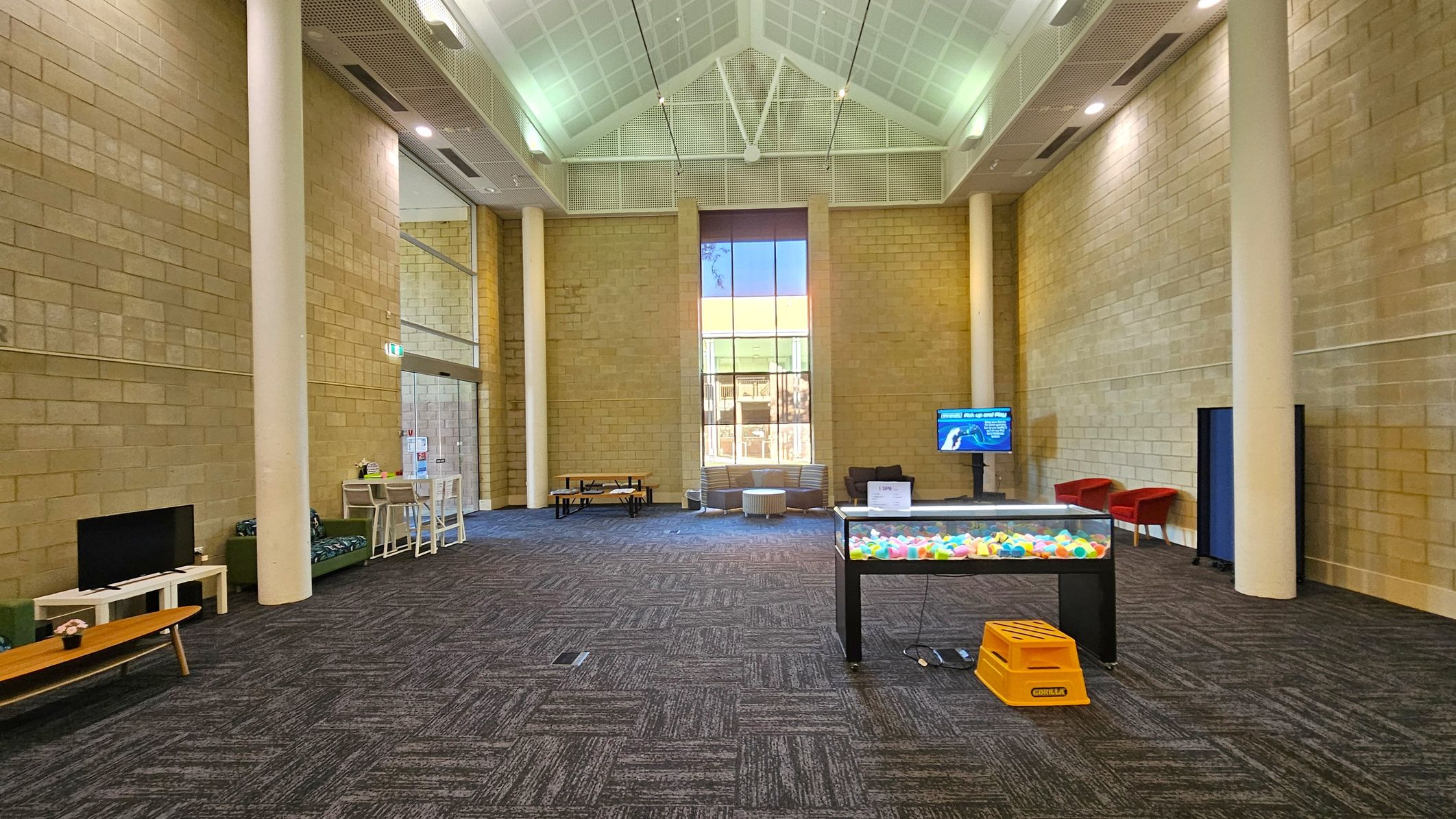 A spacious room with high ceilings and a large window in the centre background. The floor is carpeted with a dark, patterned design. On the left, there's a TV on a low table and a small seating area with a bench and chairs. In the middle, a table filled with colourful balls. On the right, a screen and two red chairs. The walls are made of light-coloured bricks, and there are tall white columns throughout the space.