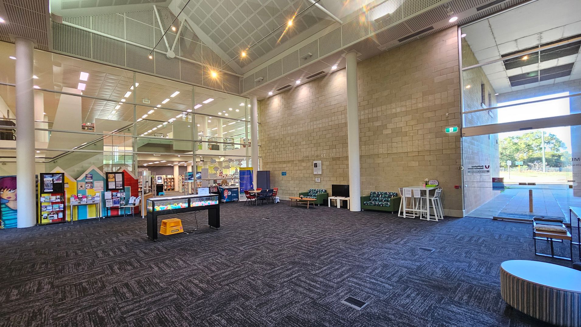 A spacious interior with a high ceiling and large glass windows. On the left, there are colourful display boards and a glimpse of a library area with bookshelves. In the centre, a table with a yellow stool and a small aquarium-like display. On the right, seating arrangements with patterned sofas and a high table with stools. The floor is carpeted with a geometric pattern, and the ceiling features a grid design with spotlights. A glass door leads outside, showing a view of trees.