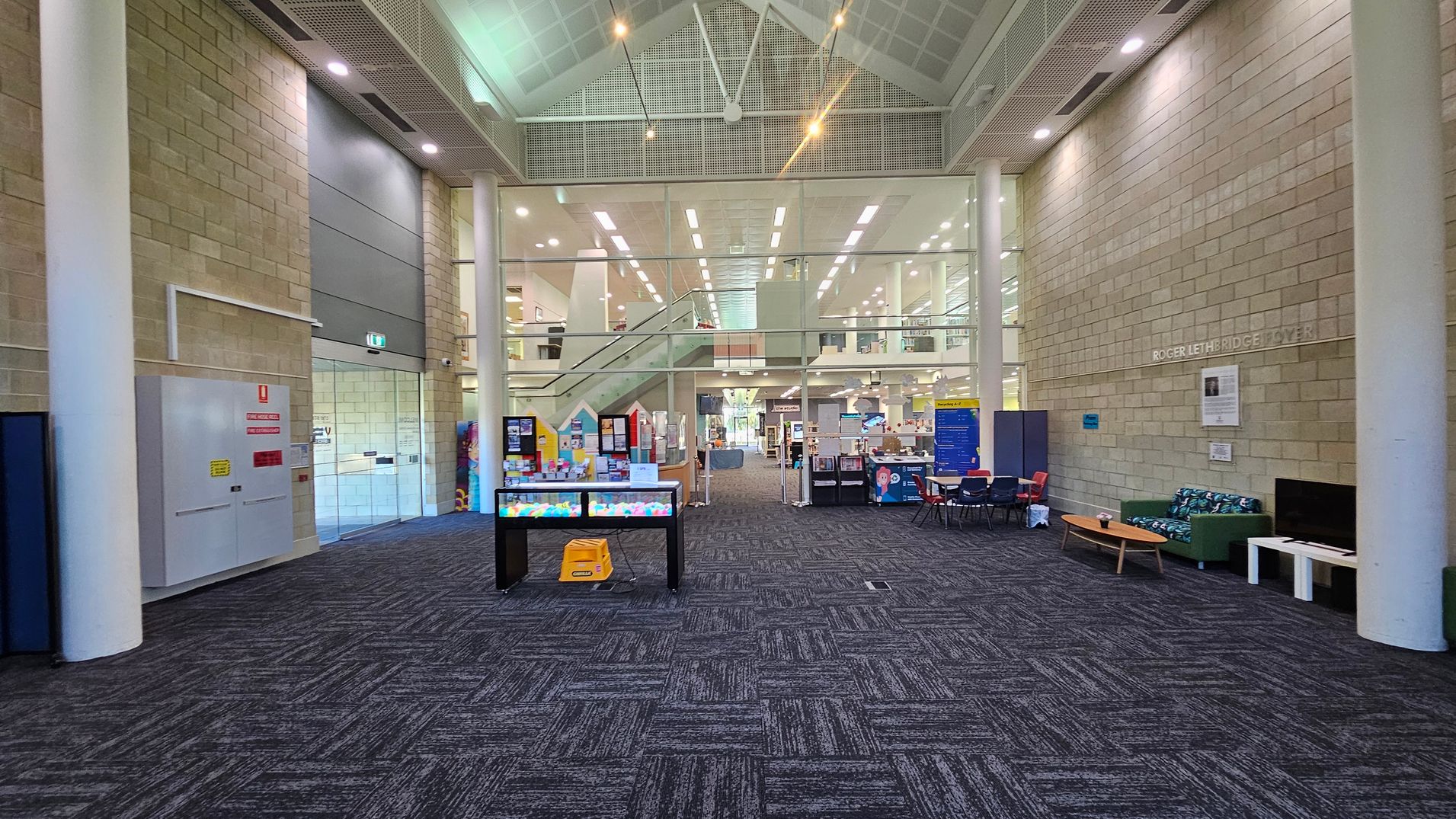 A spacious interior with a high ceiling and large glass windows. In the centre, a staircase leads to an upper level. The floor is carpeted with a dark pattern. On the left, there is a fire safety panel and a glass door. On the right, a seating area with a green sofa and a wooden coffee table is visible. Bright overhead lights illuminate the space, and various informational displays and tables are scattered throughout.