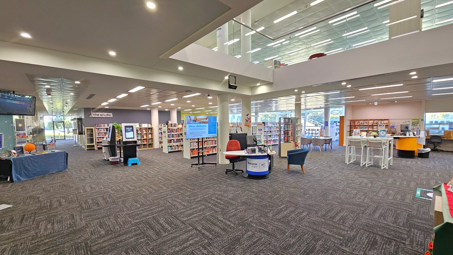 A spacious library interior with a patterned carpet floor. On the left, there is a table with a blue cloth and a pumpkin, and a TV screen mounted above. In the centre, a digital kiosk and a welcome desk with a red chair are visible. Bookshelves line the back wall, and a sign reads "the studio." On the right, there are more bookshelves, a counter with stools, and a computer station. The ceiling is lined with fluorescent lights, and large windows allow natural light in.
