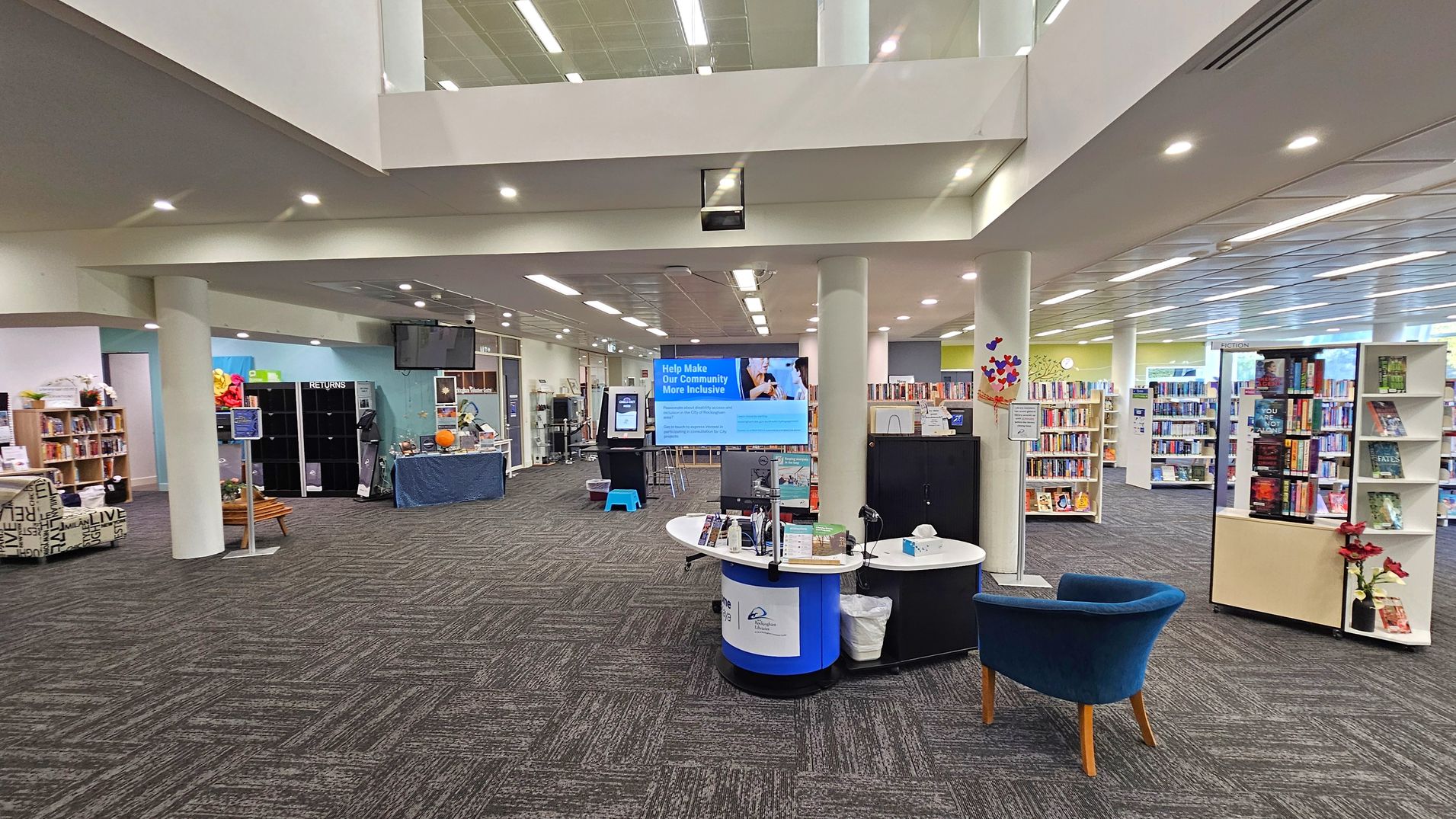 A spacious library interior with a modern design. In the foreground, a blue chair and a circular information desk with brochures and a computer. To the right, bookshelves filled with books and a small plant. In the centre, a large digital screen displaying community information. The left side features a returns area with cubbies and a table covered with a blue cloth. The ceiling is high with recessed lighting, and the floor is carpeted with a geometric pattern.