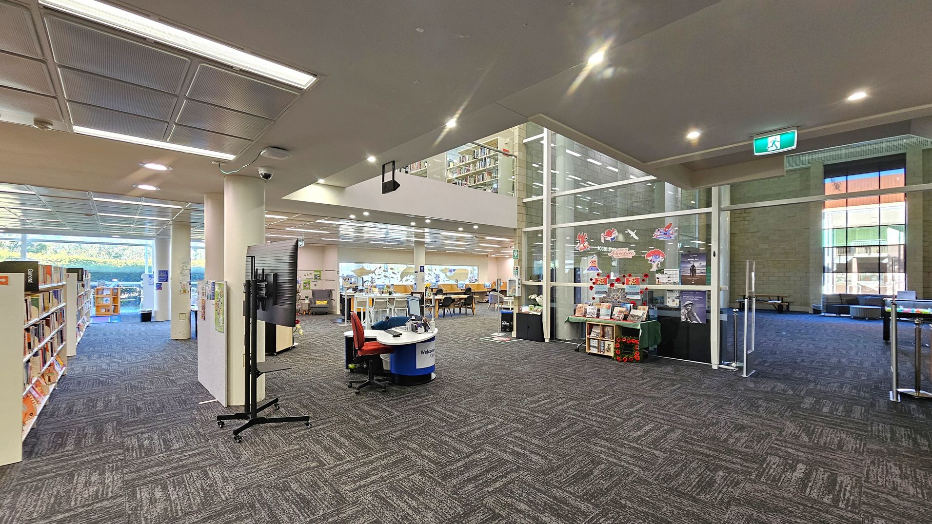 A spacious library interior with a patterned carpet floor. On the left, there are bookshelves filled with books. In the centre, a circular welcome desk with a red chair and a large screen on a stand. The right side features a glass wall with decorations and a display of books. Beyond the glass, there are more seating areas. The ceiling has recessed lighting and a visible exit sign. Large windows in the background allow natural light to illuminate the space.