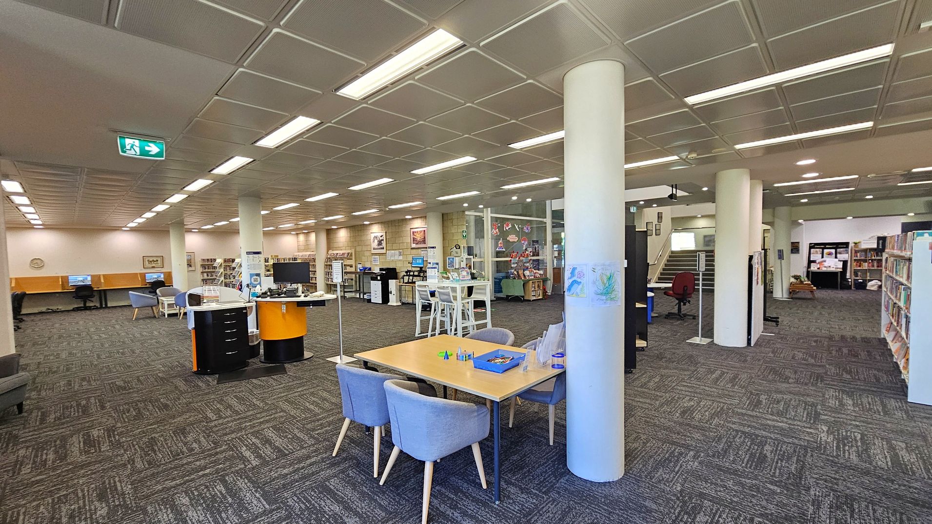 A spacious library interior with a patterned carpet floor and a grid-like ceiling with fluorescent lights. On the left, there are computer stations and bookshelves. In the centre, a round desk with a computer and a table with chairs. To the right, more bookshelves and a staircase leading upwards. A white column stands near the centre, and there are various informational signs and decorations throughout the space.