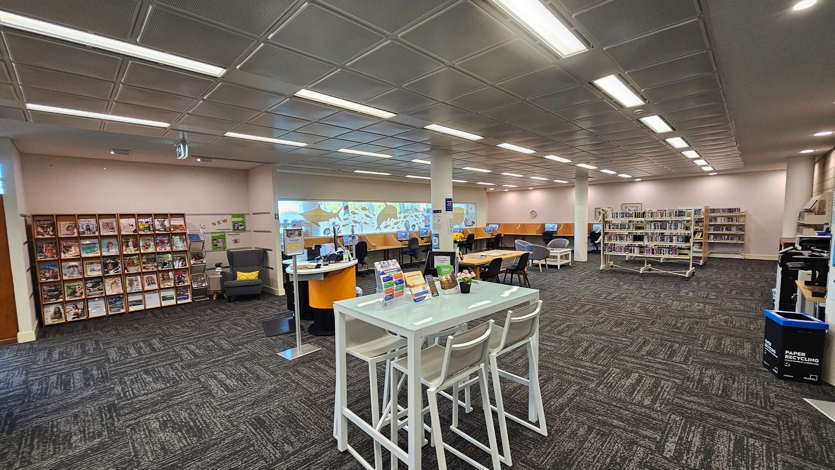 A library interior with a modern design. On the left, there is a wall with shelves filled with magazines and a comfortable armchair. In the centre, a high table with stools displays brochures. Behind it, a curved desk with computers and chairs is visible. On the right, there are bookshelves filled with books and more computer stations. The floor is carpeted with a patterned design, and the ceiling has recessed lighting panels.