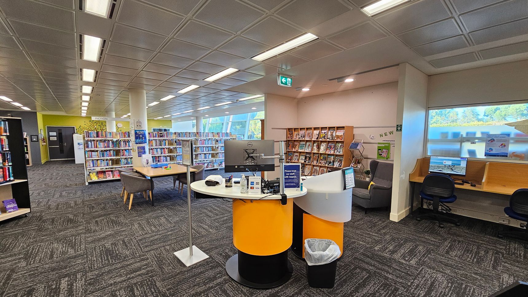 A library interior with a patterned carpet floor and a grid-patterned ceiling with fluorescent lights. On the left, there are bookshelves labelled "Fiction" and a seating area with chairs around a table. In the centre, a circular desk with a computer, hand sanitiser, and a sign. On the right, a window with a view outside and a computer workstation with chairs. Bookshelves and a display for newspapers are in the background.