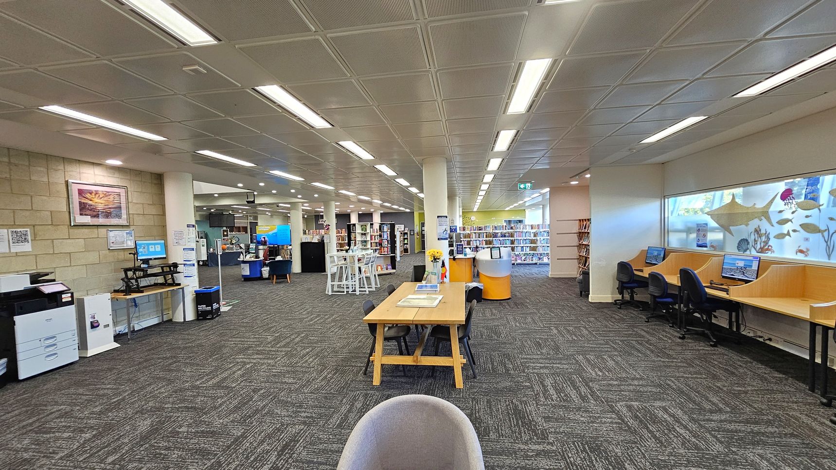 A spacious library interior with a grey carpeted floor and a grid-patterned ceiling with fluorescent lights. On the left, there's a printer and a computer station against a brick wall with framed artwork. In the centre, a wooden table with chairs and a vase of flowers. The right side features computer workstations with chairs, set against a window decorated with marine-themed decals. Bookshelves and additional seating are visible in the background.
