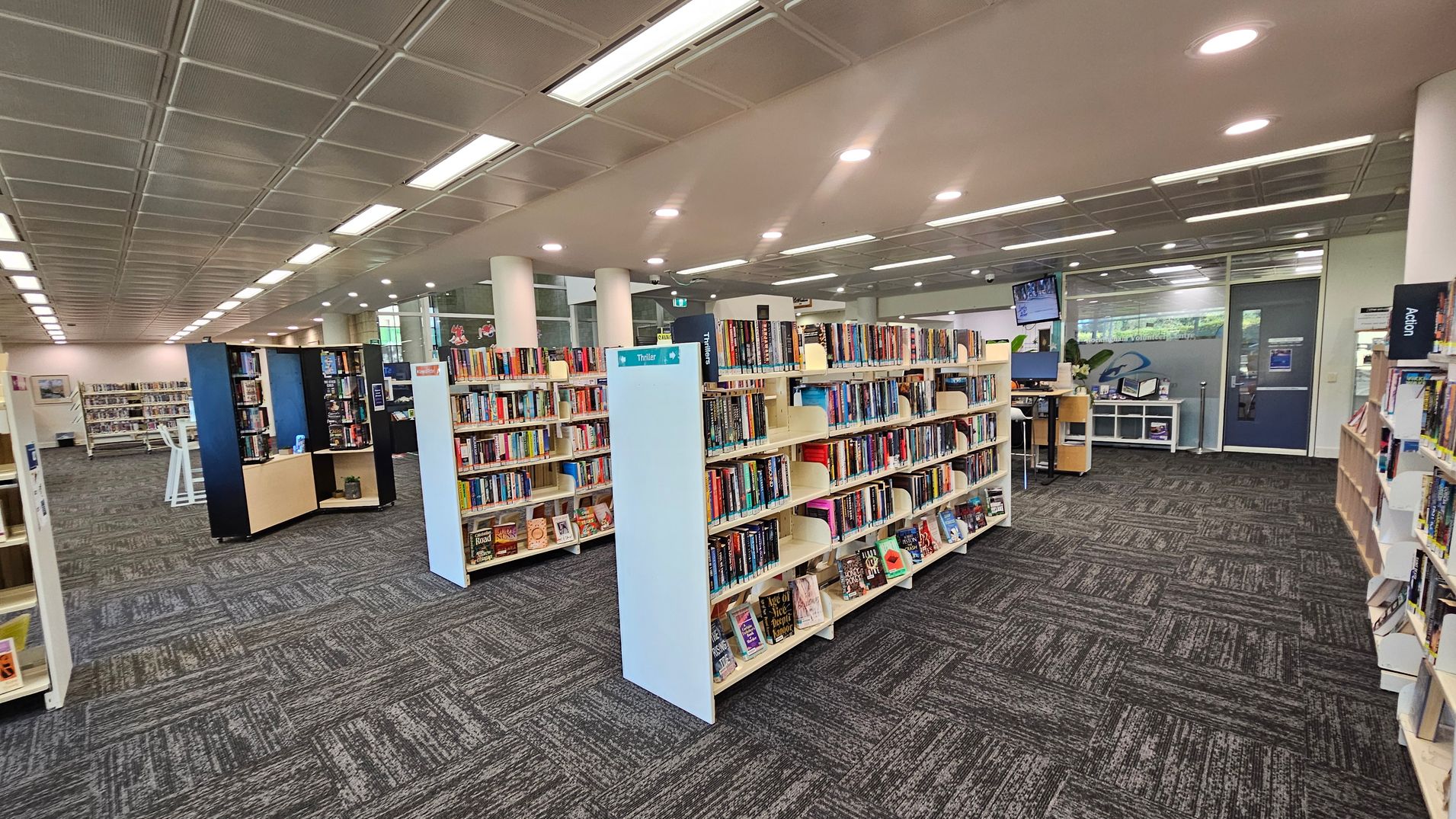 A library interior with rows of bookshelves filled with books. In the centre, a sign labelled "Thriller" is visible on a shelf. To the right, there is a desk with computers and a monitor mounted on the wall. The floor is covered with patterned carpet tiles, and the ceiling features recessed lighting. In the background, large windows and glass doors allow natural light to enter the space.