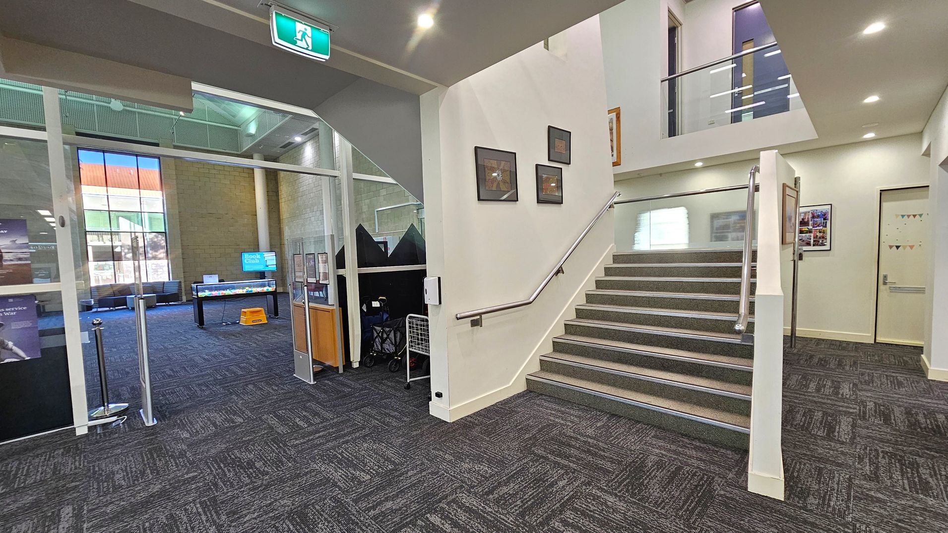 A modern interior space with a carpeted floor. On the left, there's a glass-walled room with a "Book Club" sign and seating area. In the centre, a staircase with a metal handrail leads upwards, with framed pictures on the wall beside it. On the right, a corridor with a closed door and a wall-mounted display. The ceiling has recessed lighting and an illuminated exit sign above the staircase.