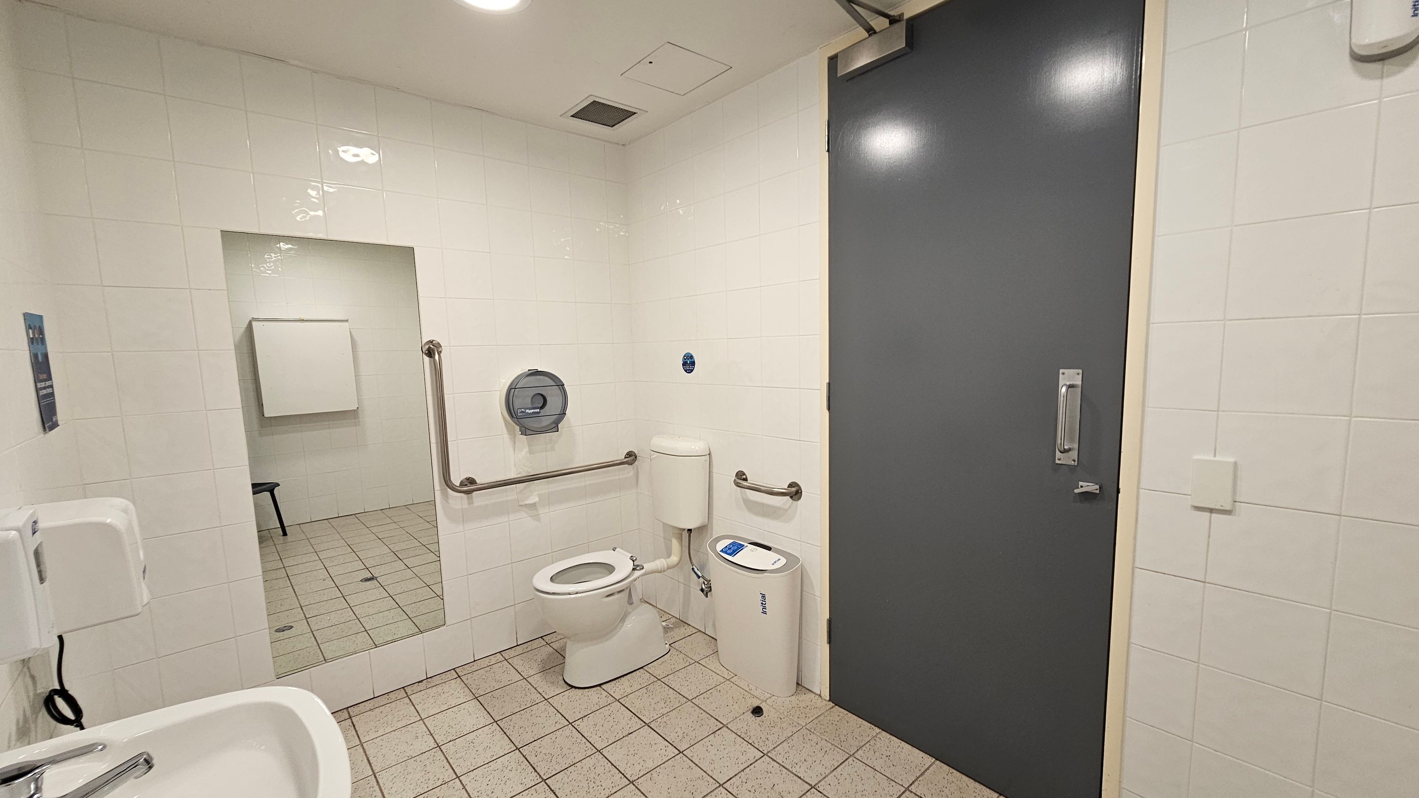 A public restroom with white tiled walls and a grey door on the right. In the centre, there is a toilet with a grab rail on each side and a toilet paper dispenser mounted on the wall. A sanitary bin is positioned next to the toilet. On the left, a sink with a soap dispenser is visible, and a large mirror reflects part of the room. The floor is covered with small square tiles, and the ceiling has recessed lighting.