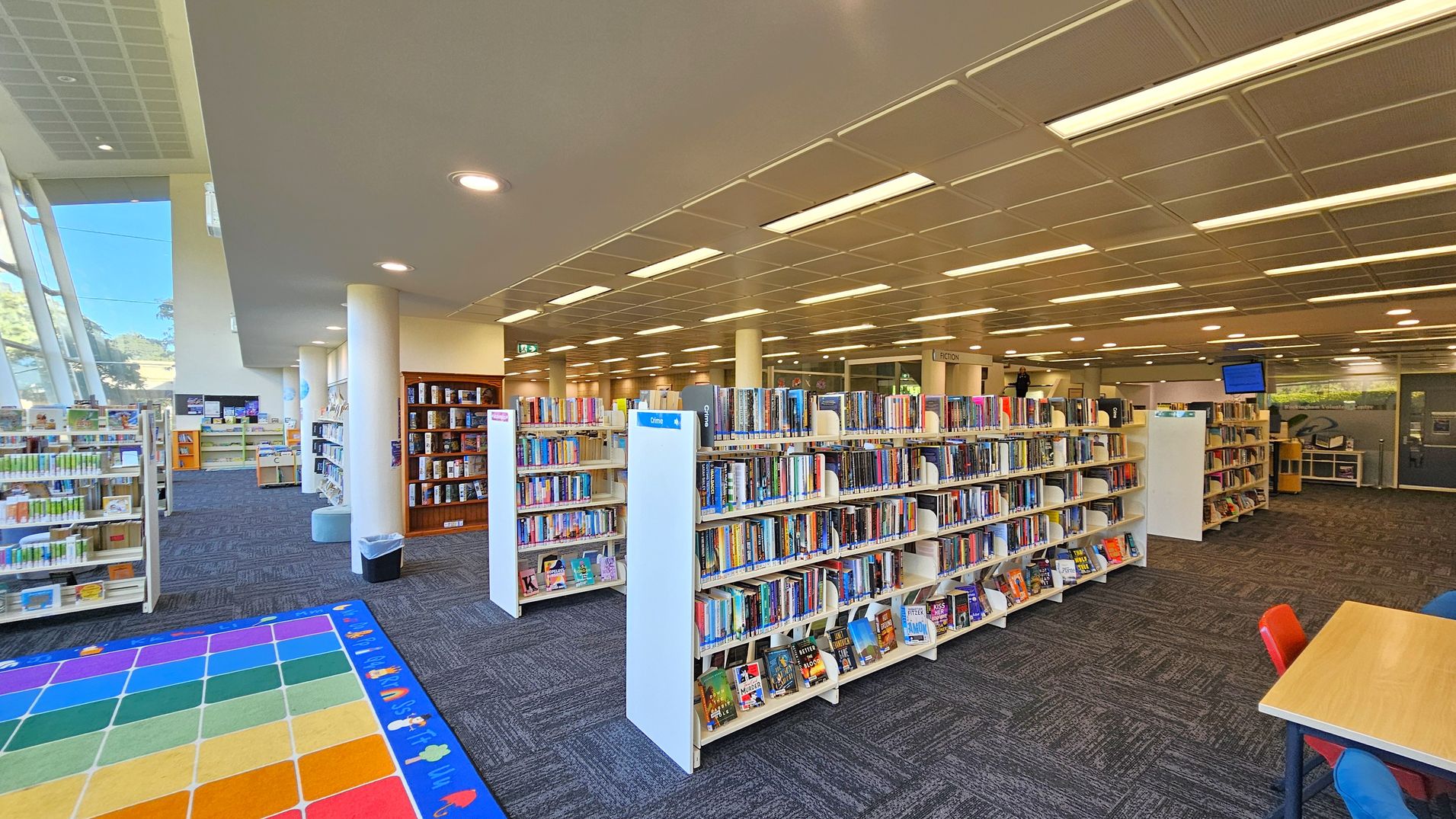 A library interior with bookshelves filled with books in the centre and right. On the left, large windows allow natural light to illuminate the space, and there are more bookshelves and a colourful children's play mat with a grid pattern. The ceiling is fitted with recessed lighting, and the floor is carpeted in a dark pattern. A table with red and blue chairs is visible in the foreground on the right.