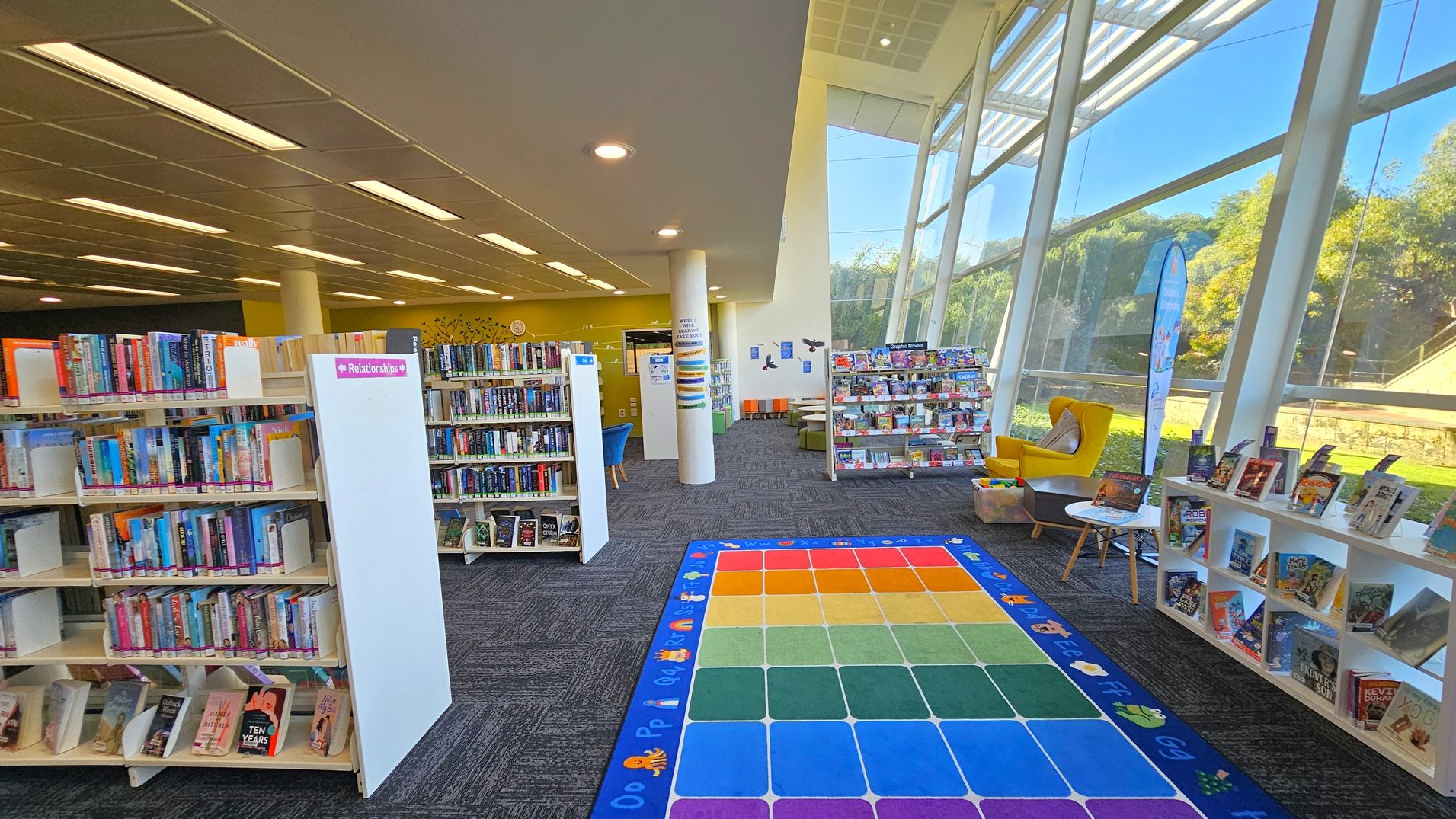 A library interior with bookshelves on the left filled with books, labelled "Relationships". In the centre, a colourful alphabet mat is on the floor. To the right, large windows allow natural light, with a yellow armchair and a small table nearby. More books are displayed on a low shelf. The ceiling has recessed lighting, and the floor is carpeted in a dark pattern. In the background, there are more bookshelves and seating areas.