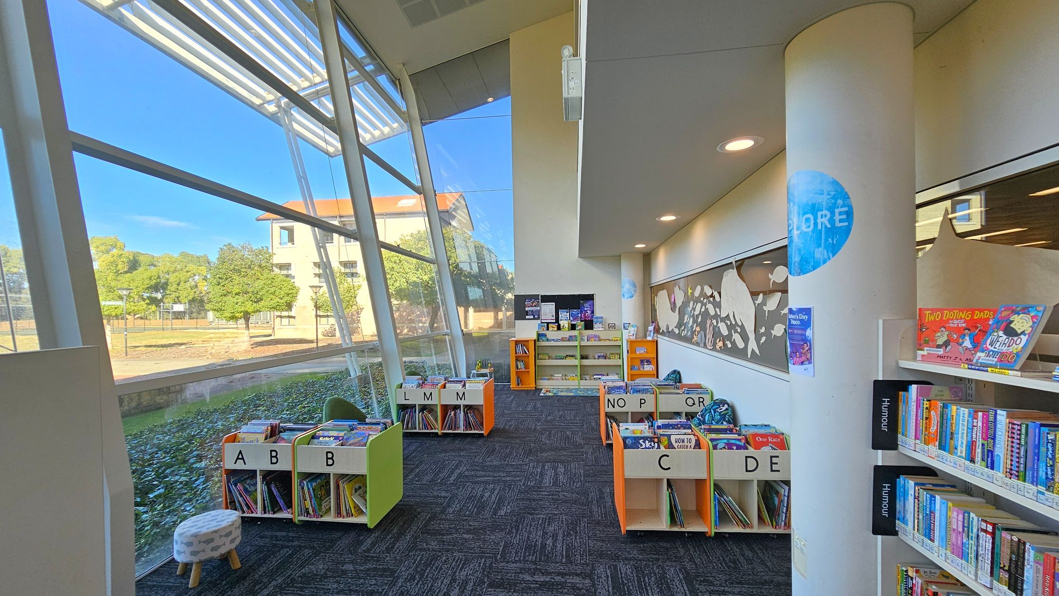 A library interior with large, slanted glass windows on the left, offering a view of trees and a building outside. In the centre, there are several orange and white book bins labelled with letters, filled with children's books. A column on the right has a blue circle with the word "EXPLORE". Bookshelves on the right hold various books, with some labelled "Humour". The floor is carpeted in a dark pattern, and the ceiling has recessed lighting.
