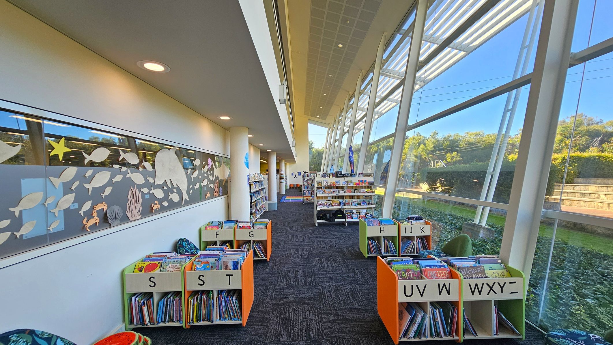 A library interior with a high ceiling and large glass windows on the right, allowing natural light to illuminate the space. On the left, a wall features a decorative panel with cut-out shapes of fish and sea creatures. The floor is carpeted in a dark pattern. In the centre, there are low bookshelves with books organised alphabetically, labelled with letters. Further back, taller bookshelves are visible, and outside the windows, greenery and a pathway can be seen.