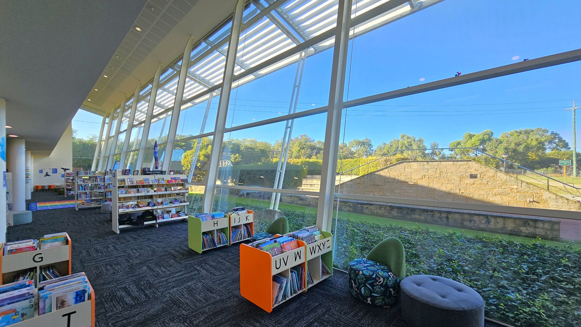 A library interior with large, slanted glass windows on the right, allowing natural light to illuminate the space. Bookshelves filled with books are arranged in the centre and left, with sections labelled by letters. The floor is carpeted in a dark pattern, and there are colourful seating options, including a round pouffe and a green chair. Outside, greenery and a stone wall are visible through the windows. The ceiling is high and features recessed lighting.