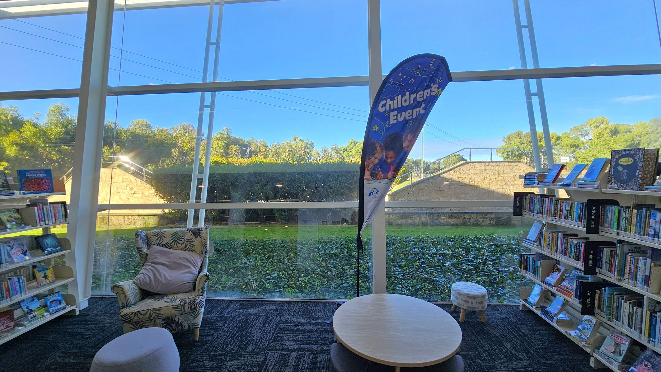 A library interior with large glass windows in the background, revealing a view of greenery and a stone wall outside. In the foreground, there is a patterned armchair with a cushion, a small round table, and a stool. To the left and right are bookshelves filled with books, with signs indicating sections like "Adventure" and "Animals." A banner stands near the window, advertising "Children's Events." The floor is carpeted in a dark pattern.