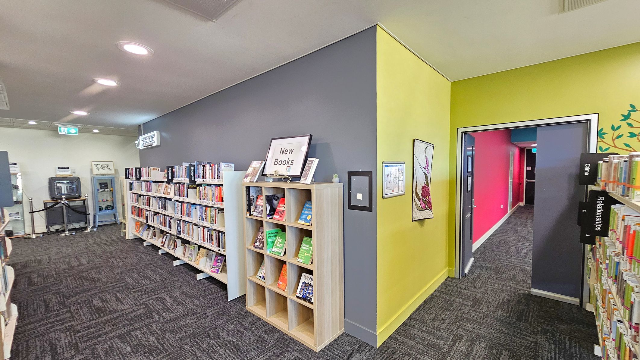 Bookshelves line the left wall, filled with books, and a sign labelled "New Books" sits atop a smaller shelf. The floor is carpeted in a dark pattern. On the right, a doorway with a bright pink wall beyond leads to another area. A sign on the right shelf indicates sections for "Crime" and "Relationships". The walls are painted grey and yellow, with a decorative piece hanging on the yellow wall. The ceiling is white with recessed lighting.