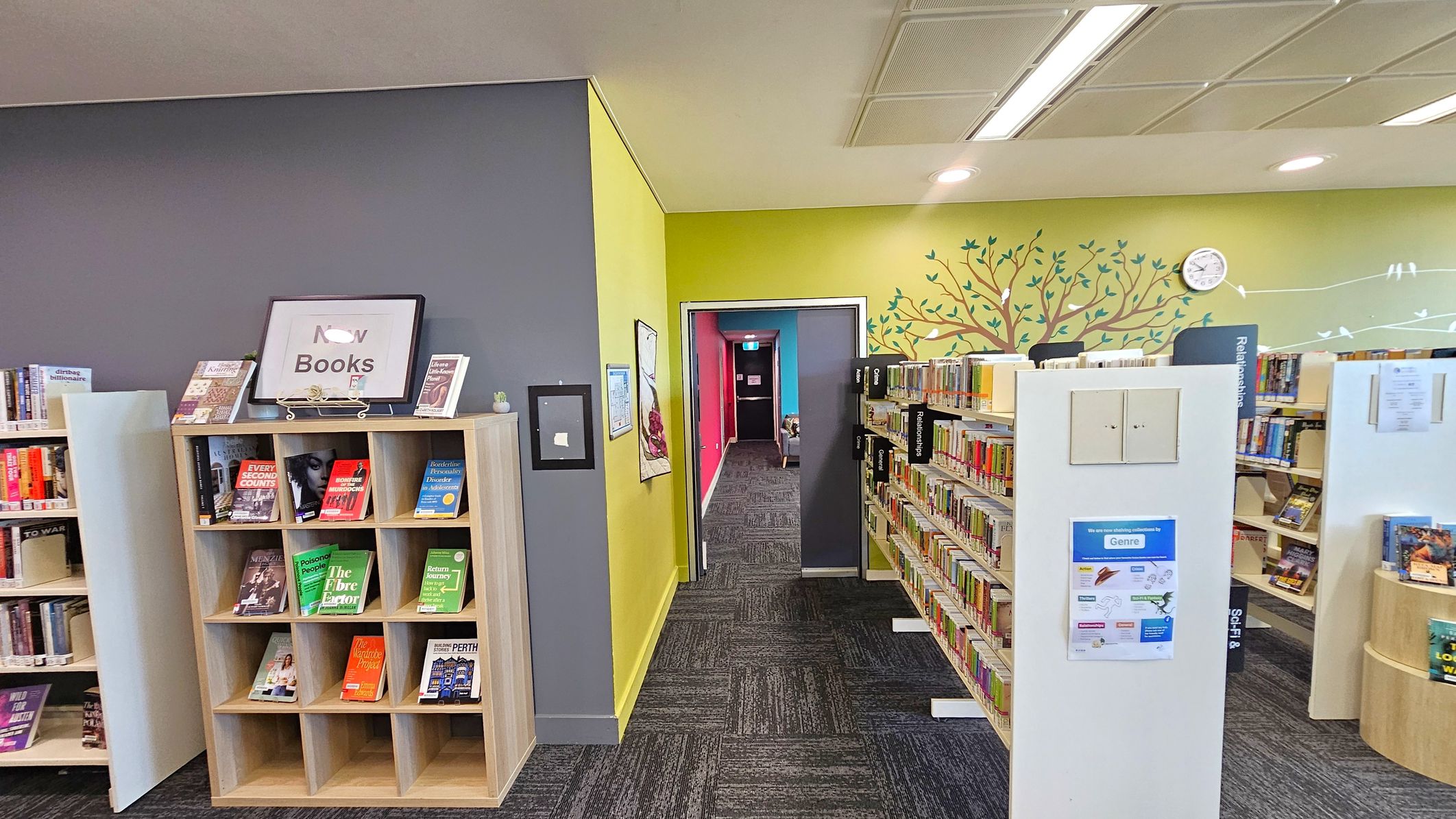 A library interior with a grey and yellow wall. On the left, a wooden bookshelf displays various books with a sign on top. In the centre, a hallway with a red and blue wall leads to another area. On the right, bookshelves are filled with books, and a wall features a tree mural with a clock. The ceiling has recessed lighting, and the floor is carpeted with a dark pattern.