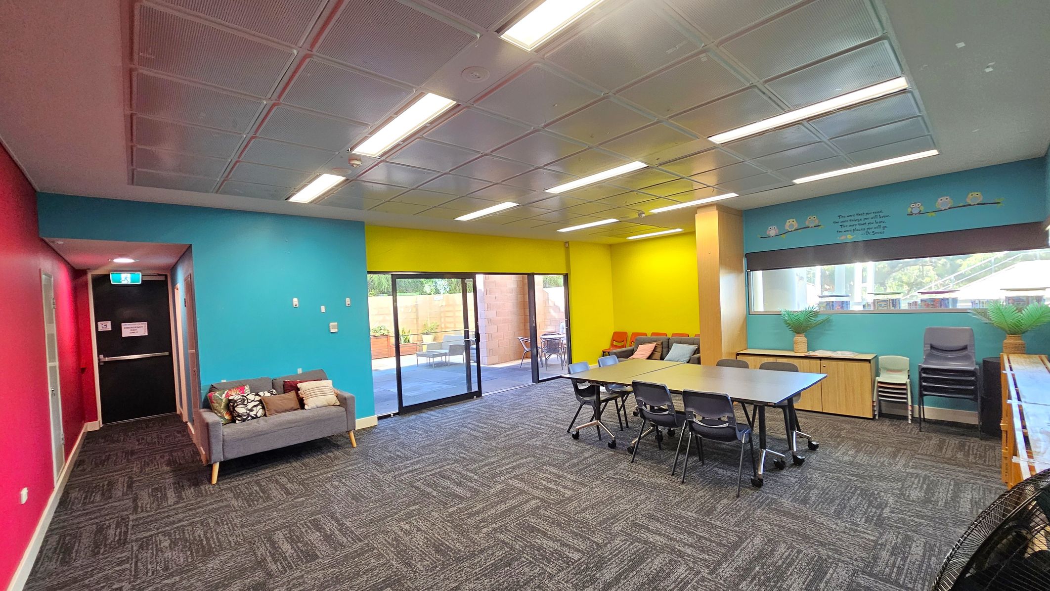 A room with vibrant walls: pink on the left, blue in the middle, and yellow on the right. A grey sofa with cushions is against the blue wall. A glass door leads to an outdoor area with chairs. A table with chairs is in the centre. The right wall has a window with a blind, potted plants, and stacked chairs. The ceiling is tiled with fluorescent lights, and the floor is carpeted with a geometric pattern.