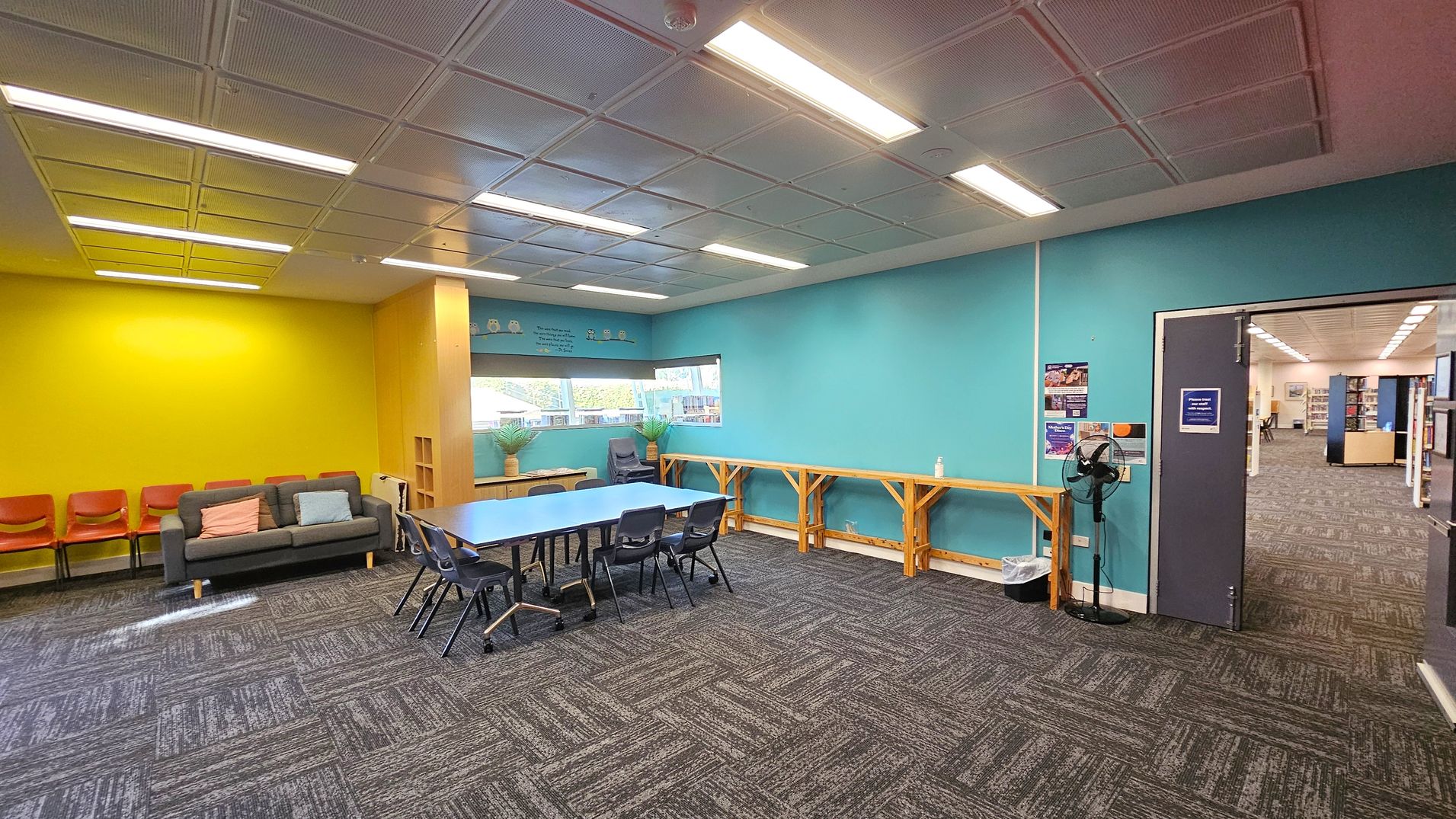 A room with a yellow wall on the left featuring a grey sofa with cushions and several orange chairs. In the centre, a blue table with black chairs is placed on a patterned carpet. The right wall is turquoise with a wooden counter and a fan. A doorway leads to another room with bookshelves. The ceiling has recessed lighting, and there are windows with potted plants on a small table. Posters are visible on the turquoise wall near the door.