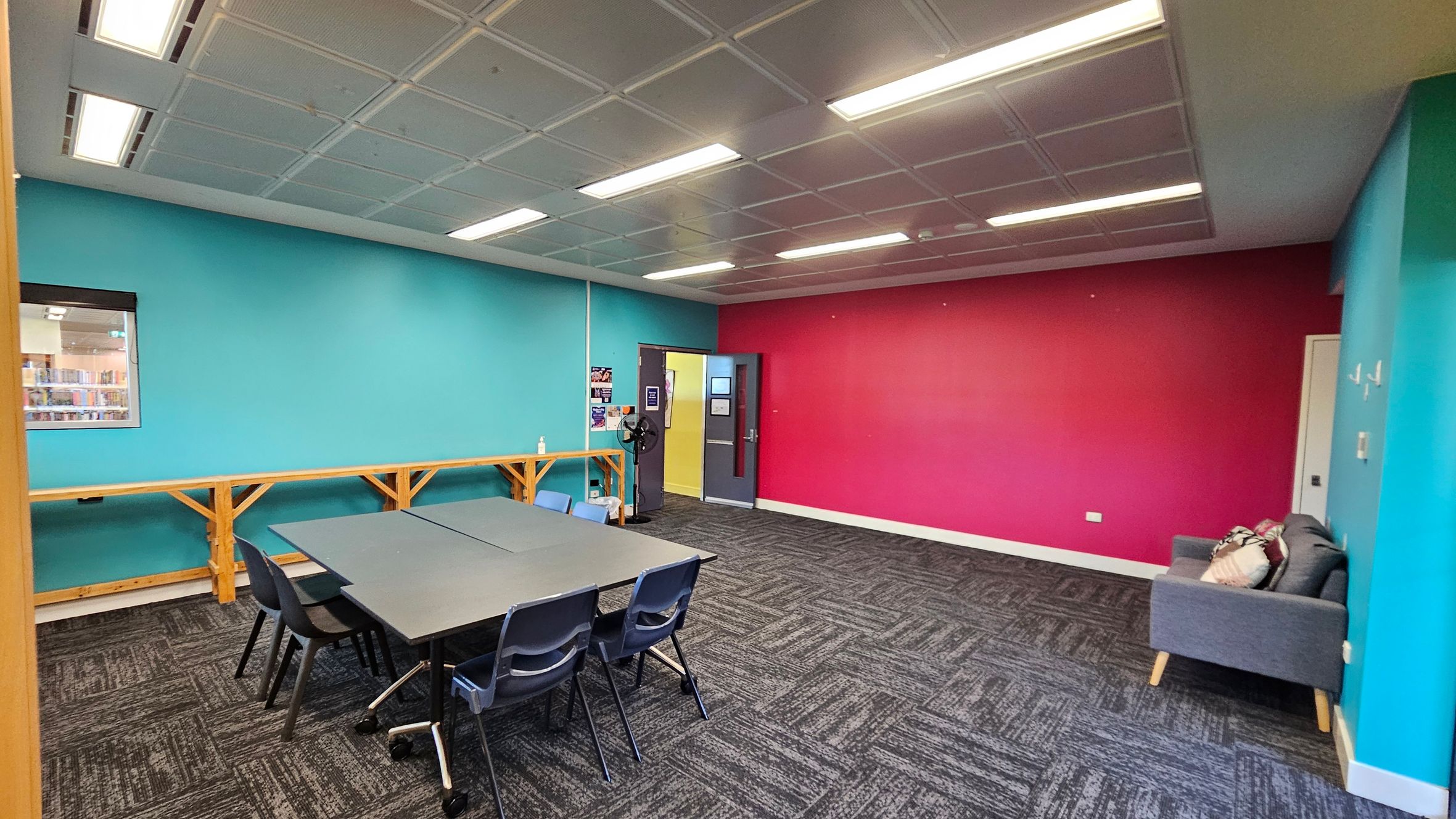 A room with a turquoise wall on the left and a bright pink wall on the right. In the centre, there is a grey table surrounded by black and blue chairs. A wooden bench runs along the turquoise wall. A grey sofa with cushions is against the pink wall. The floor is covered with dark patterned carpet tiles. The ceiling has a grid pattern with fluorescent lights. A door in the pink wall leads to another room, and a window on the left wall shows a glimpse of bookshelves.