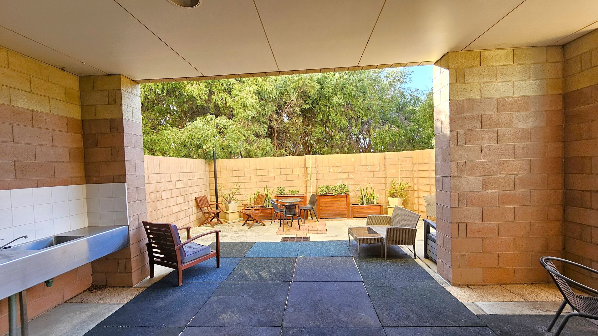 A covered outdoor area with a tiled ceiling and brick walls. On the left, there's a stainless steel sink and a wooden chair. The floor is covered with dark rubber tiles. In the centre, an open patio features a round table with chairs, surrounded by potted plants and wooden planters. On the right, there are more chairs and a small table. The background shows a brick wall with trees visible above it.