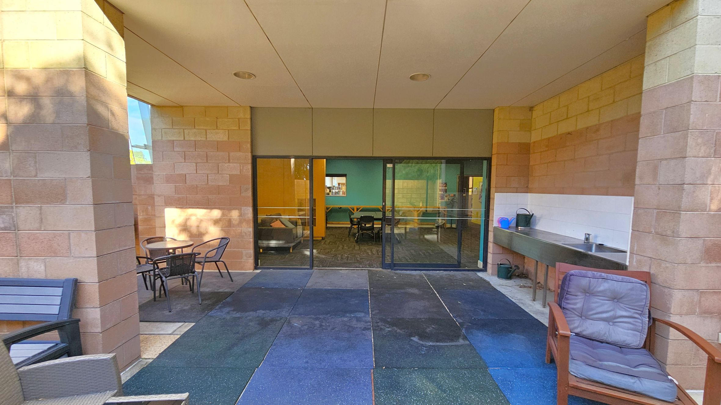 A covered outdoor area with a tiled floor and a ceiling with recessed lights. On the left, there are two round tables with chairs. On the right, a cushioned wooden chair is next to a stainless steel sink with a countertop, where a watering can and a blue container are placed. In the centre, glass doors lead to an indoor space with visible furniture, including a sofa and chairs. The walls are made of light-coloured bricks.