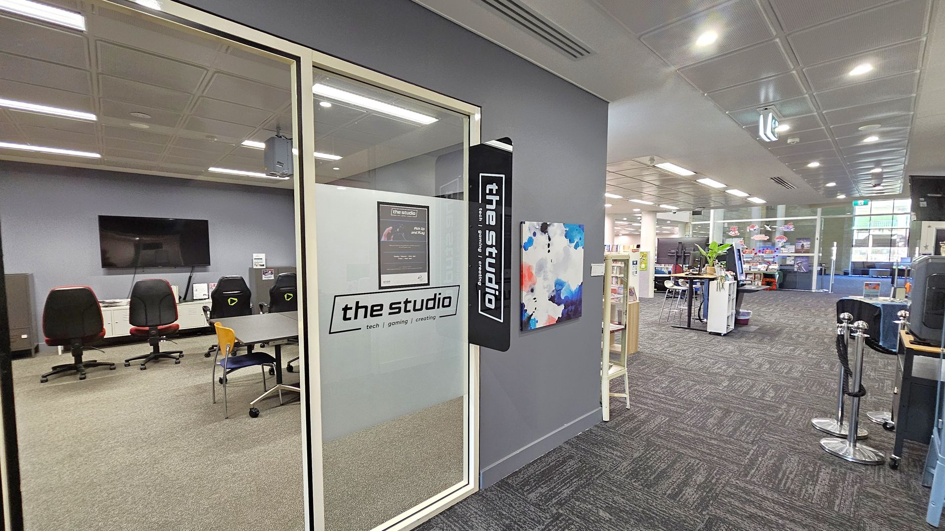 A modern interior space with a glass-walled room on the left labelled "the studio" featuring tech, gaming, and creating. Inside are several gaming chairs and a large screen. To the right, the open area has bookshelves, a high table with stools, and a plant. The floor is carpeted with a dark pattern, and the ceiling has recessed lighting. A colourful abstract painting is mounted on the wall near the studio entrance.