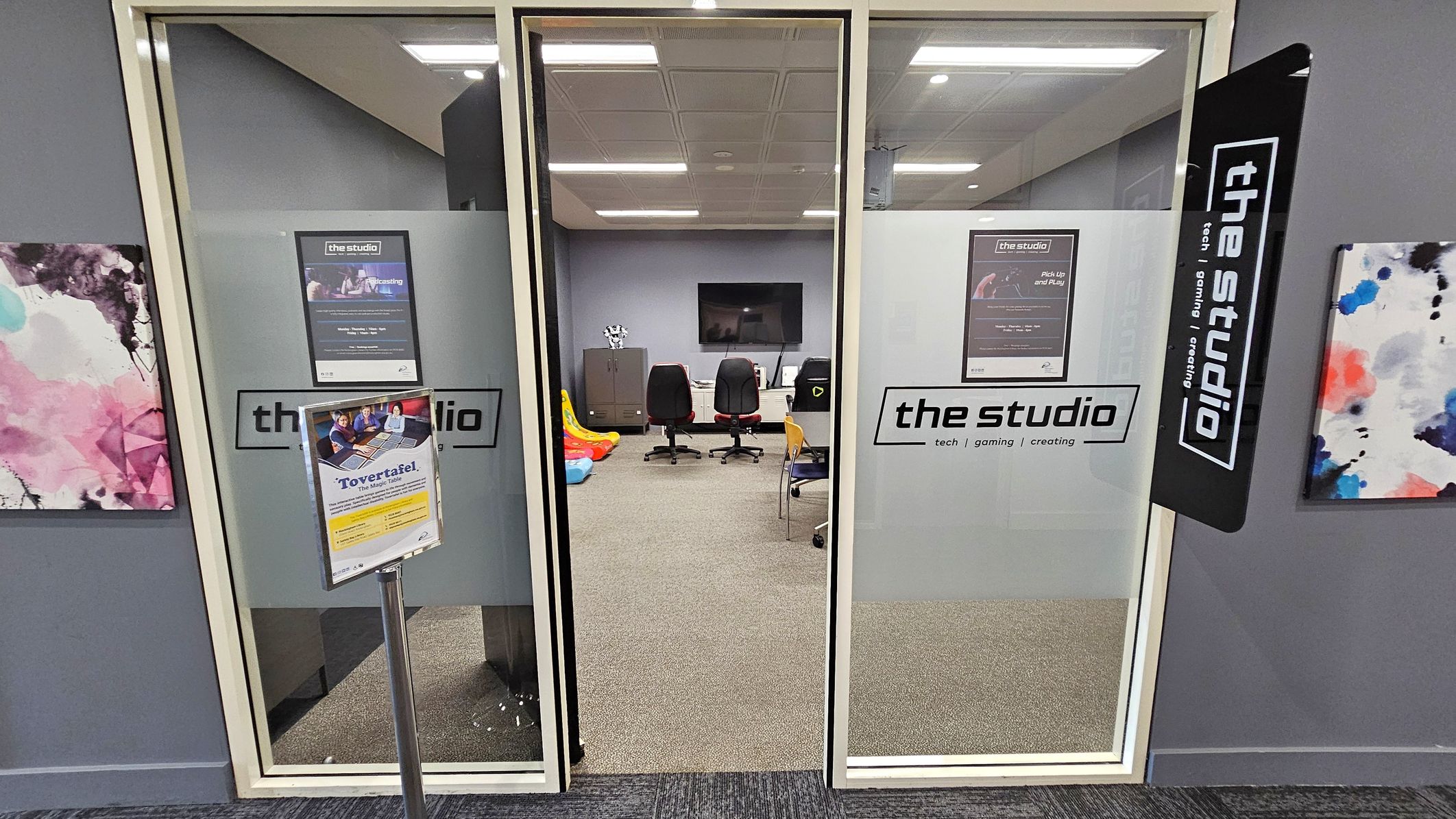 A glass entrance with two doors labelled "the studio" leads into a room. Inside, there are three office chairs facing a wall-mounted screen. To the left, colourful inflatable toys are visible. A sign on a stand is in front of the doors, and abstract art pieces hang on the walls. The ceiling has recessed lighting, and the floor is carpeted.
