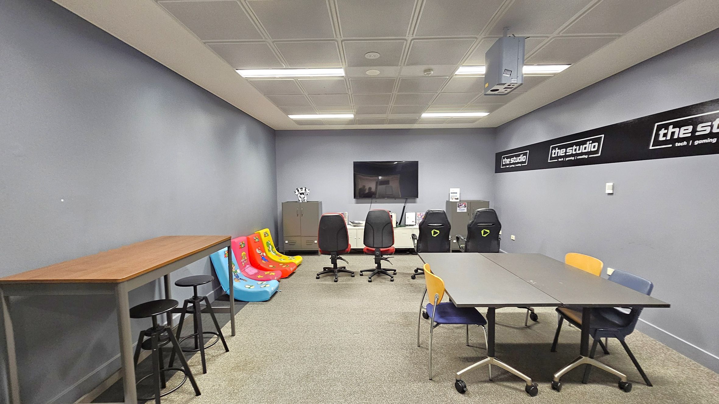 A room with grey walls and a tiled ceiling with fluorescent lights. On the left, there's a long wooden table with black stools. In the middle, colourful floor chairs are placed against the wall, with a row of gaming chairs in front of a wall-mounted TV. Cabinets are beneath the TV. On the right, a table with wheels is surrounded by colourful chairs. The floor is carpeted, and there are signs on the walls that read "the studio" with additional text.