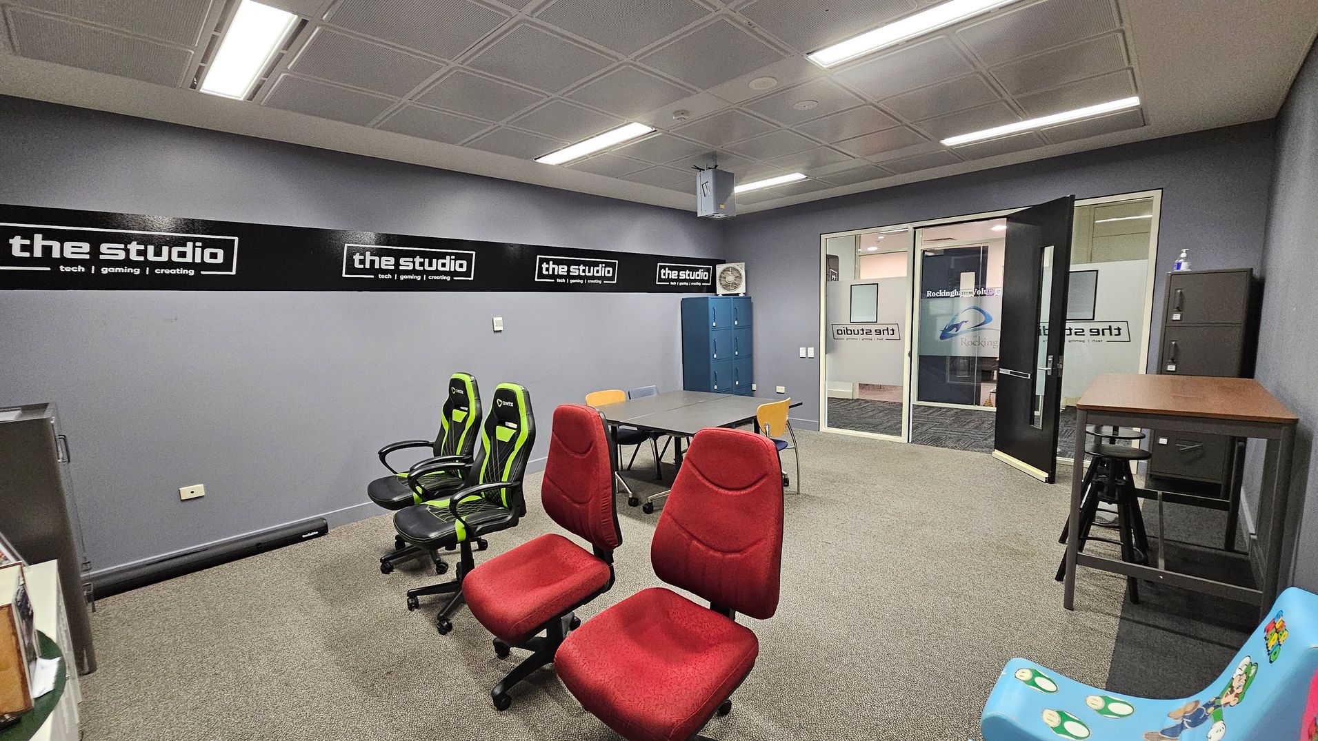 A room with grey walls and a patterned ceiling with fluorescent lights. On the left, there are two green and black gaming chairs and two red office chairs. In the centre, a table with assorted chairs. On the right, a high table with a stool and a filing cabinet. A blue chair with cartoon characters is in the foreground. The back wall has a sign reading "the studio" and a glass door with more signage. The floor is carpeted in a neutral tone.