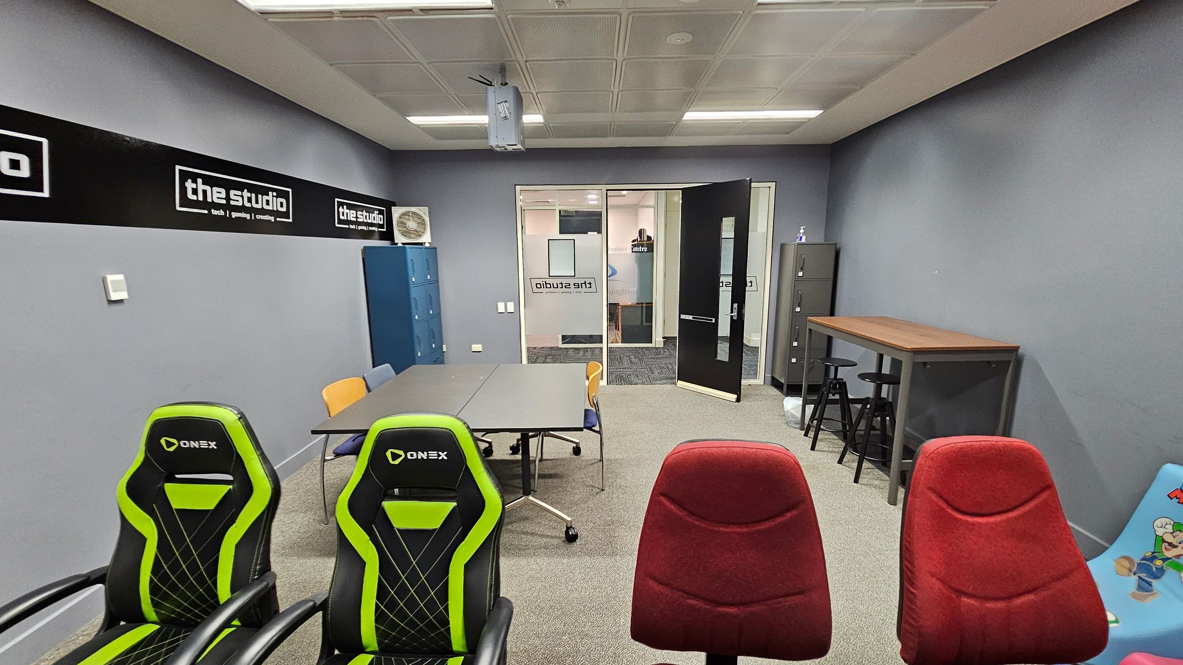 A room with grey walls and a carpeted floor. In the foreground, there are two green and black gaming chairs and two red chairs. In the middle, a grey table with assorted chairs. On the left, blue lockers and a wall-mounted air conditioner. On the right, a tall table with black stools and a colourful children's chair. The back wall has a glass door with "the studio" signage, leading to another room. The ceiling has fluorescent lights and a mounted projector.
