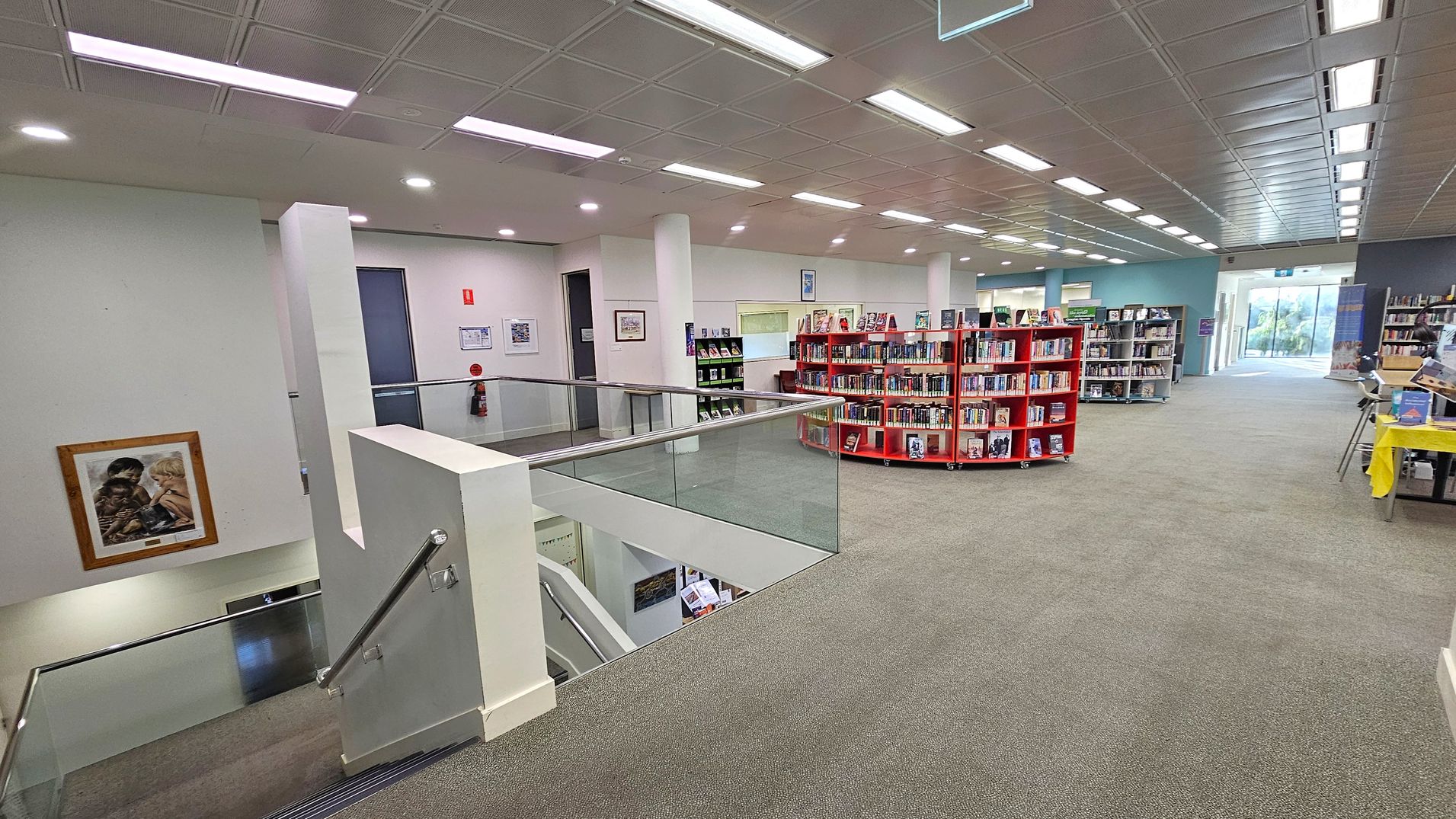 A library interior with a green carpeted floor. On the left, a staircase with a metal railing leads downwards, and a framed picture hangs on the wall. In the centre, red bookshelves are filled with books. The right side features more bookshelves and a table with a yellow cloth. The ceiling has a grid pattern with fluorescent lights. In the background, large windows allow natural light to enter.