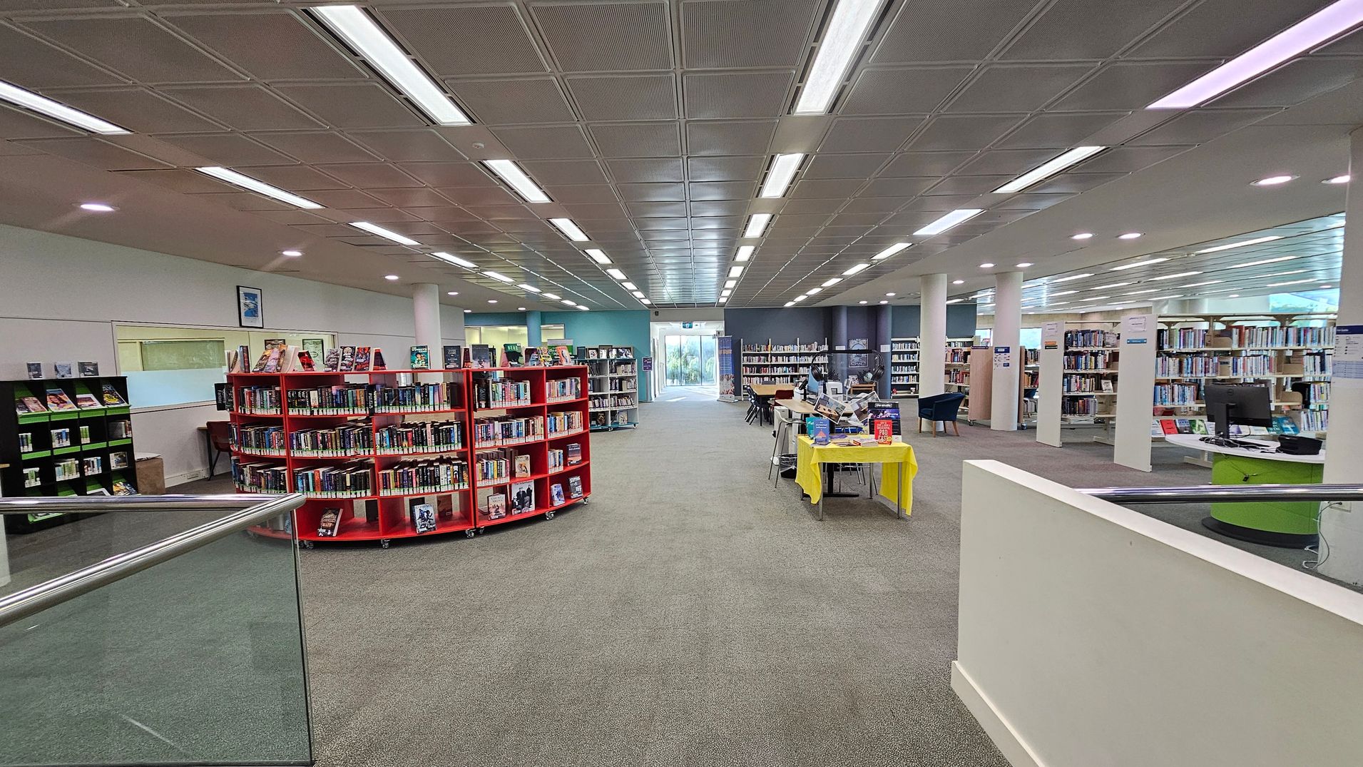 A spacious library interior with a grey carpeted floor. In the centre, there are red circular bookshelves filled with books. To the right, a yellow table displays more books, and a computer desk is visible. The left side has additional bookshelves and a framed picture on the wall. The ceiling is lined with fluorescent lights, and large windows at the far end let in natural light.