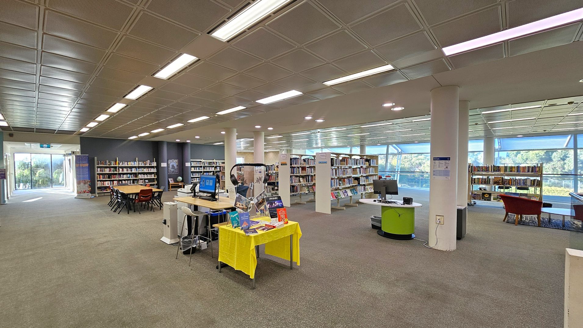 A spacious library interior with a grey carpeted floor and a ceiling with rectangular light panels. On the left, there are bookshelves against a dark wall and a large windowed exit. A wooden table with chairs is in the centre, surrounded by more bookshelves. In the foreground, a small table with a yellow cloth displays books. On the right, a circular desk with computers and a red chair near a window overlooking greenery.