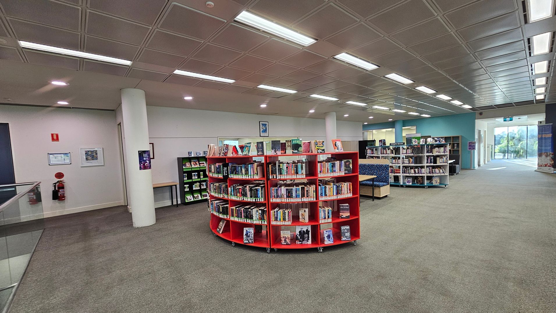 A library interior with a green carpeted floor and a ceiling with recessed lighting. In the centre, there is a circular red bookshelf filled with books. To the right, more bookshelves and a seating area with blue cushioned benches are visible. On the left, a white wall with a fire extinguisher and framed notices. The background shows a glass entrance with natural light coming in, and a blue wall with additional bookshelves.
