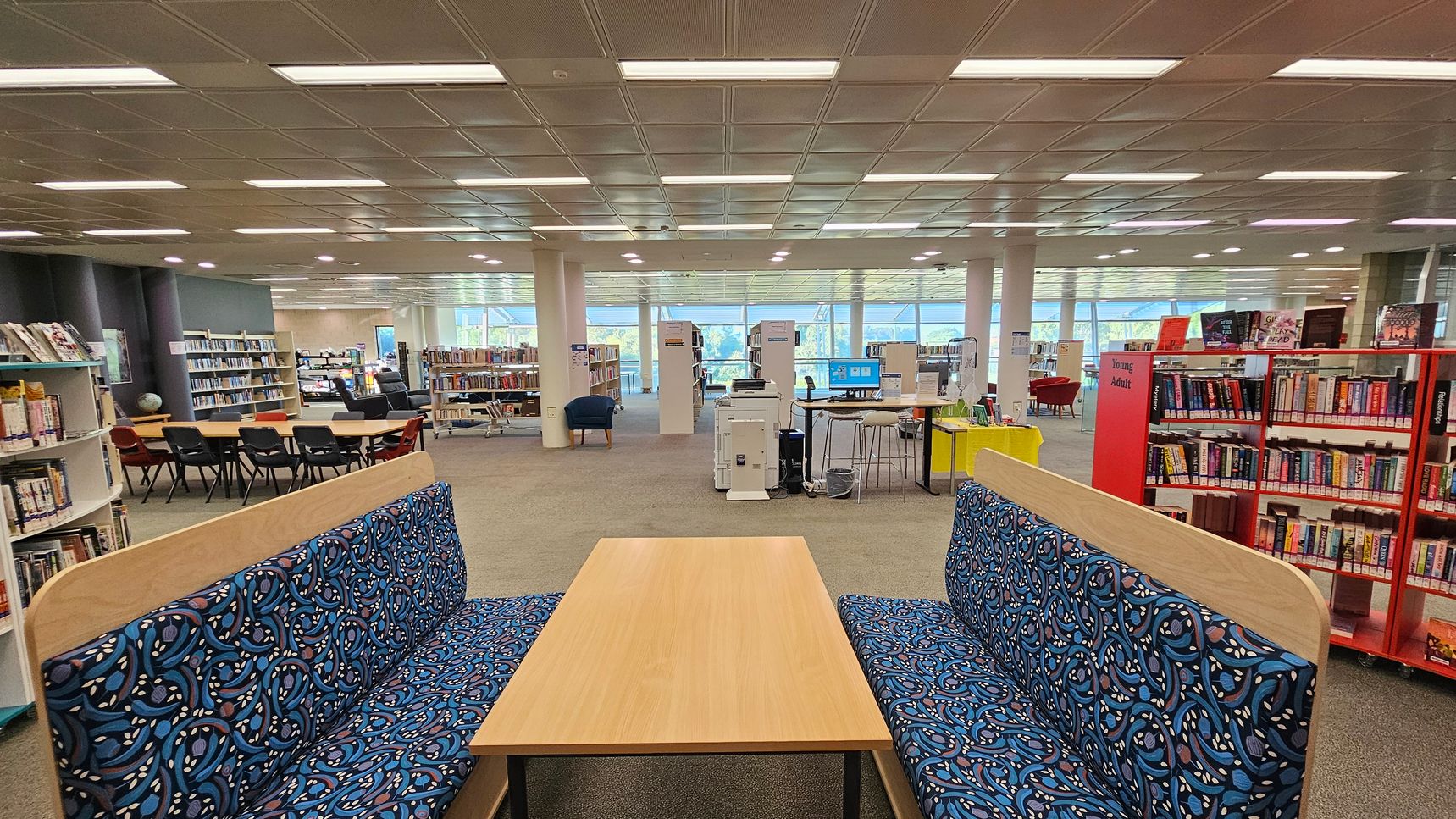 A library interior with a central seating area featuring a wooden table and two benches with blue patterned upholstery. To the left, there are bookshelves and a round table with chairs. On the right, red bookshelves labelled "Young Adult" are visible. In the background, there are more bookshelves, a computer station, and large windows allowing natural light. The ceiling is lined with fluorescent lights, and the floor is carpeted.
