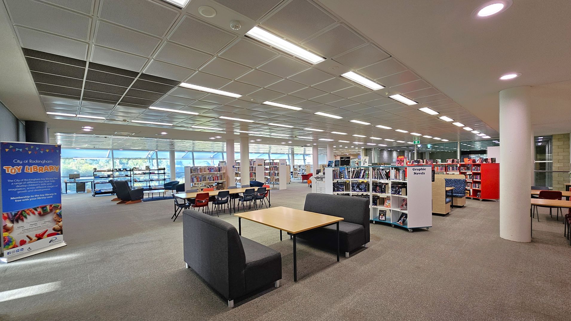 A spacious library interior with a grey carpeted floor and a grid-patterned ceiling with fluorescent lights. On the left, a banner for the "City of Rockingham Toy Library" stands near large windows. In the centre, there are tables with chairs and a seating area with two grey sofas facing a wooden table. On the right, bookshelves labelled "Graphic Novels" are filled with books. The background features more bookshelves and seating areas, with natural light streaming through the windows.