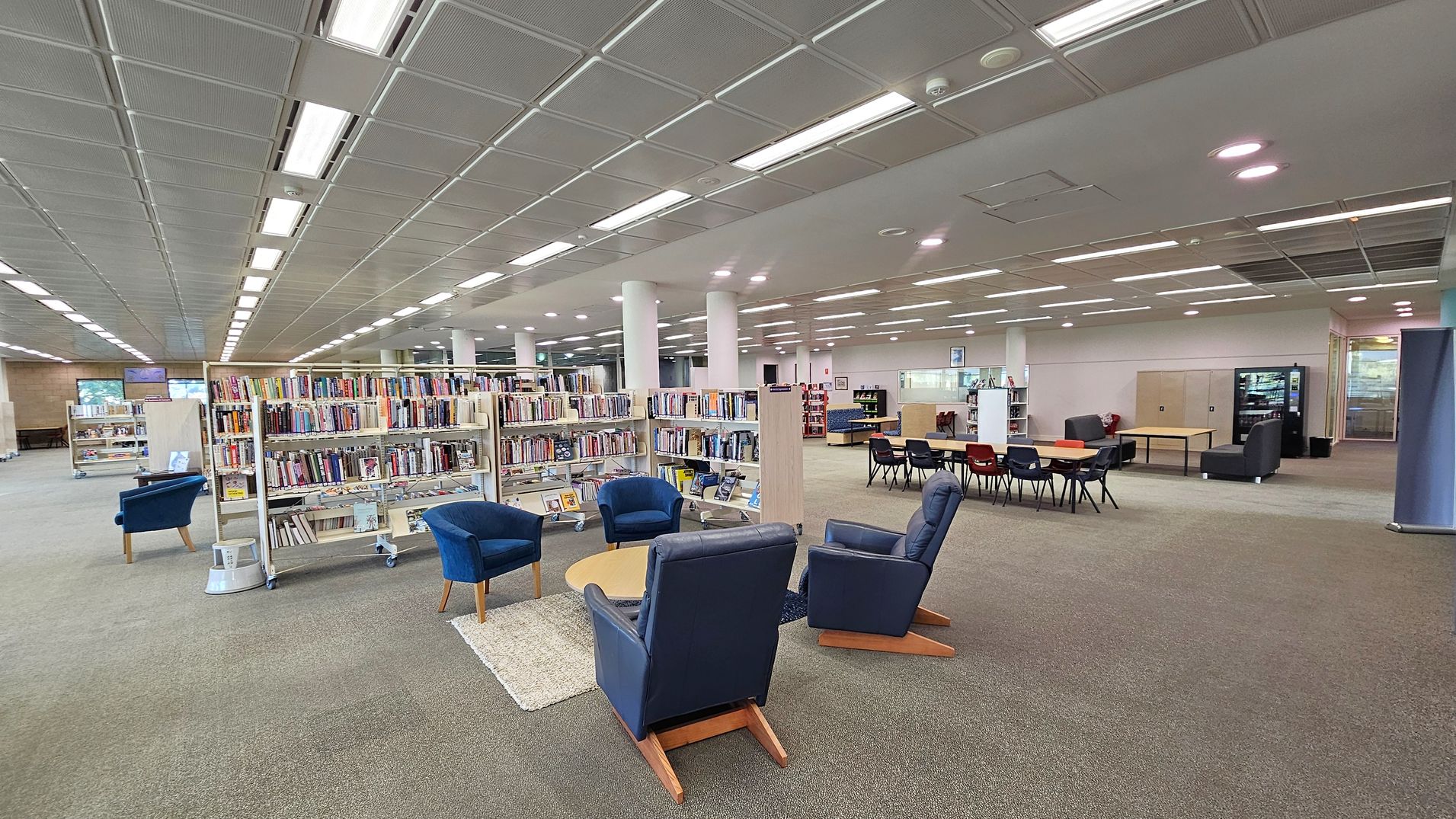 A spacious library interior with bookshelves filled with books in the centre. On the left, there are blue armchairs around a small round table on a rug. To the right, a seating area with tables and chairs, and a vending machine in the background. The floor is carpeted, and the ceiling features a grid pattern with fluorescent lighting. White columns are evenly spaced throughout the room.