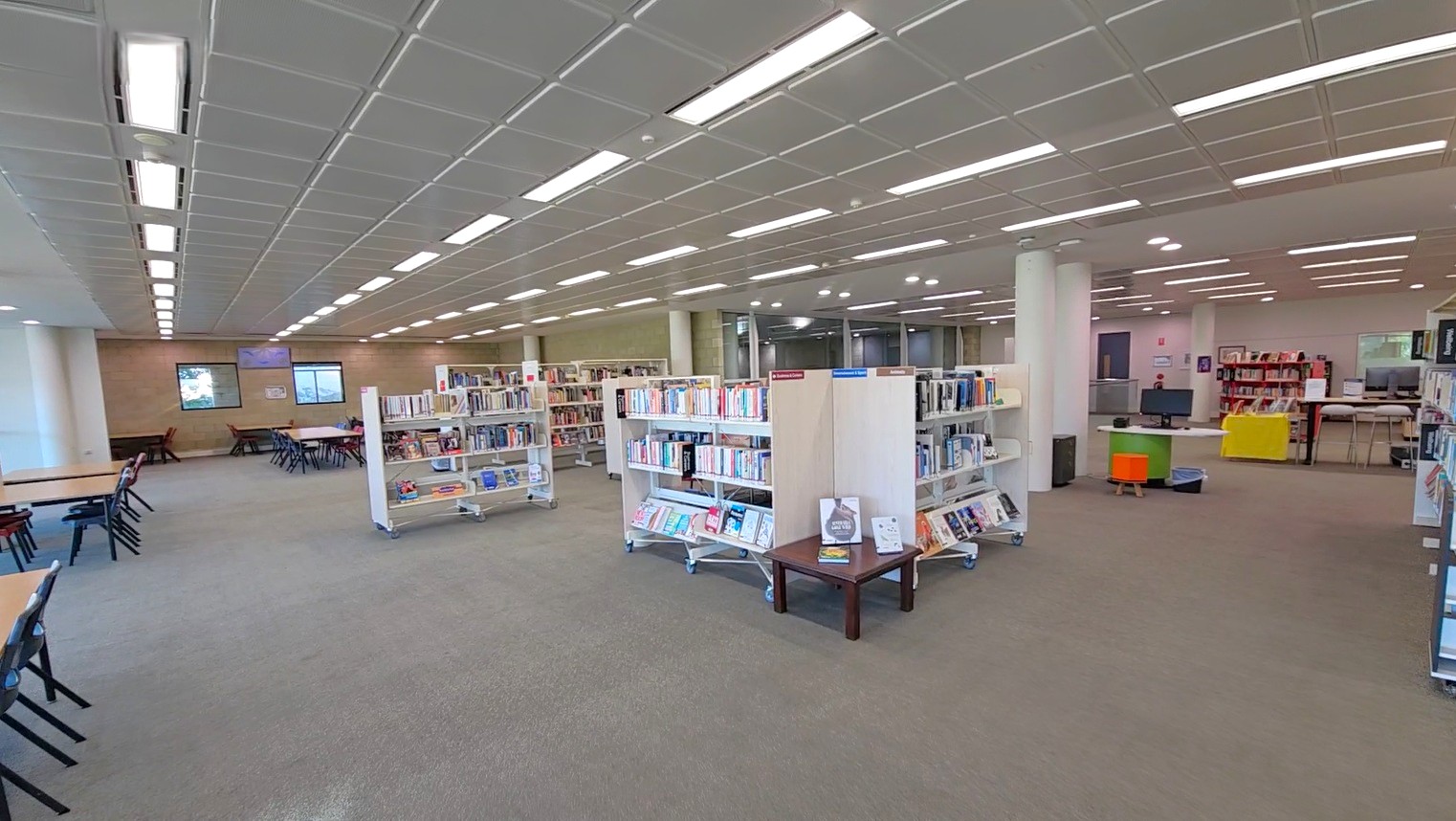 A spacious library interior with a grey carpeted floor. In the foreground, several white bookshelves filled with books are arranged in rows. To the left, there are tables with chairs for reading or studying. The ceiling is lined with rectangular fluorescent lights. In the background, more bookshelves and a few colourful chairs are visible, along with a computer station. The walls are light-coloured, and there are windows on the left side.