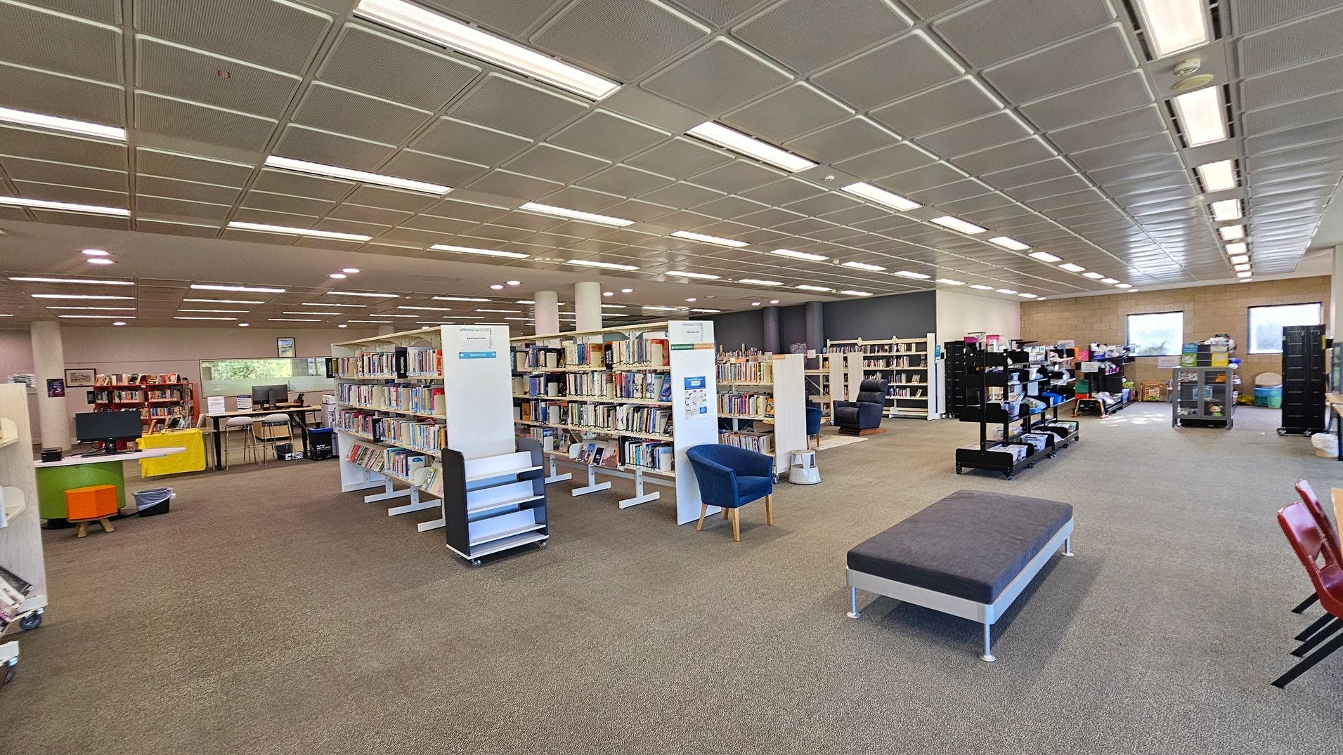 A spacious library interior with a grey carpeted floor and a ceiling with rectangular light panels. In the centre, there are several white bookshelves filled with books, accompanied by blue chairs. On the left, there are computer stations with orange and green stools. To the right, there are black shelves with various items and two windows in the background. A grey bench and red chairs are visible in the foreground.