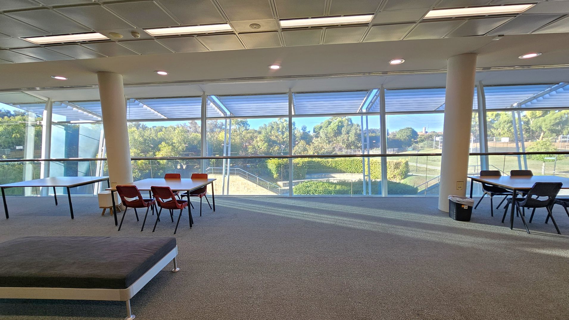 A spacious room with large floor-to-ceiling windows in the background, offering a view of greenery outside. On the left, there are tables with red chairs, and on the right, more tables with black chairs. The floor is carpeted in grey, and the ceiling has recessed lighting and a grid pattern. Two white columns are visible, supporting the structure.