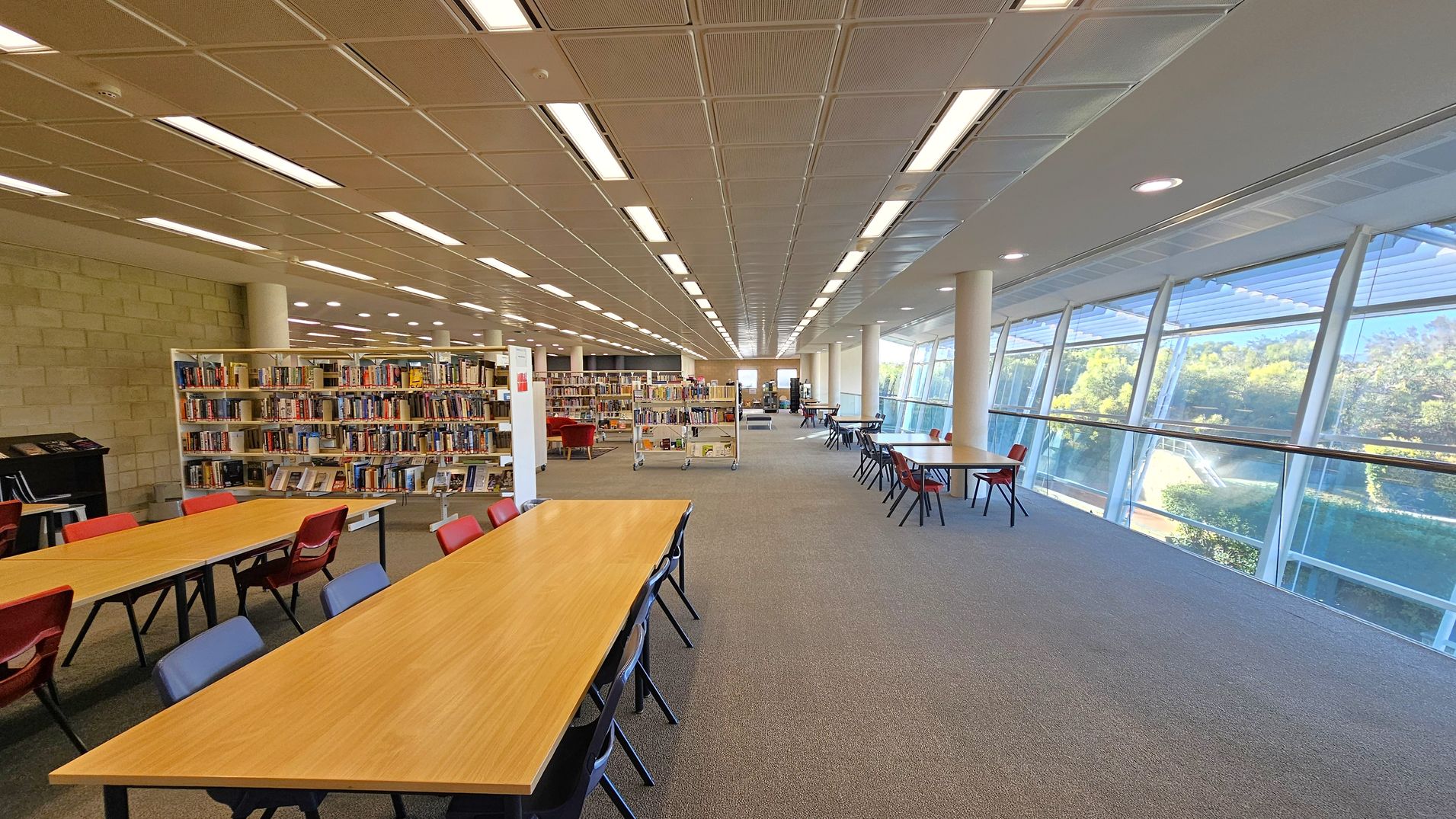 A spacious library interior with a long row of wooden tables and red chairs on the left. Bookshelves filled with books are positioned centrally. On the right, large windows provide a view of greenery outside, allowing natural light to flood in. The floor is carpeted in grey, and the ceiling features a grid pattern with fluorescent lighting.
