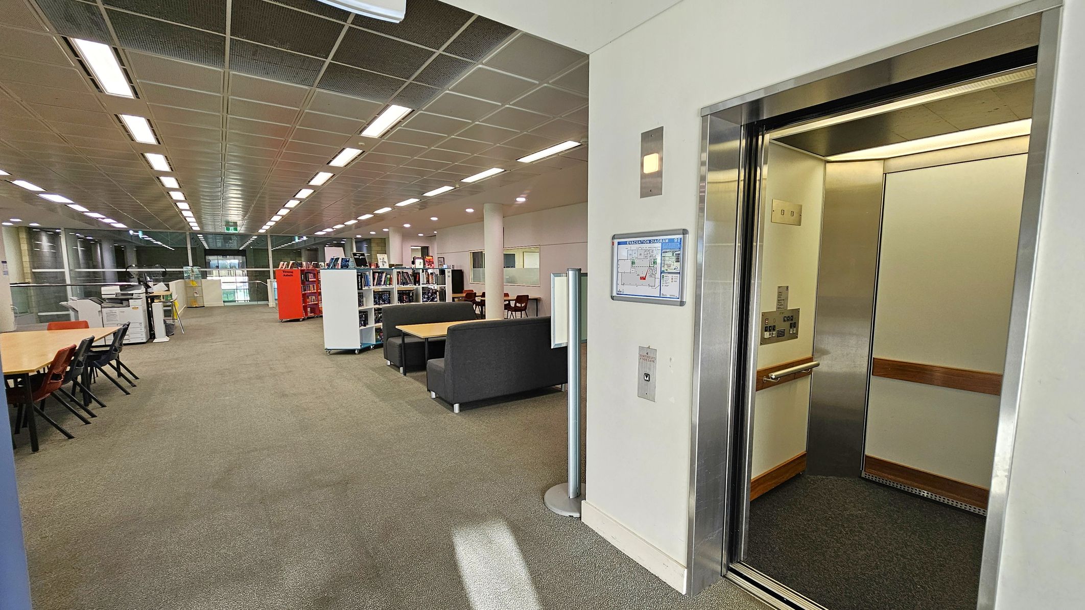 An open lift is on the right with a metallic interior and wooden handrails. A floor plan is mounted on the wall beside the lift. The left side features a spacious area with tables, chairs, and bookshelves. There are sofas and a copier further back. The ceiling has a grid pattern with fluorescent lights, and the floor is carpeted. Large windows are visible in the background, providing natural light.