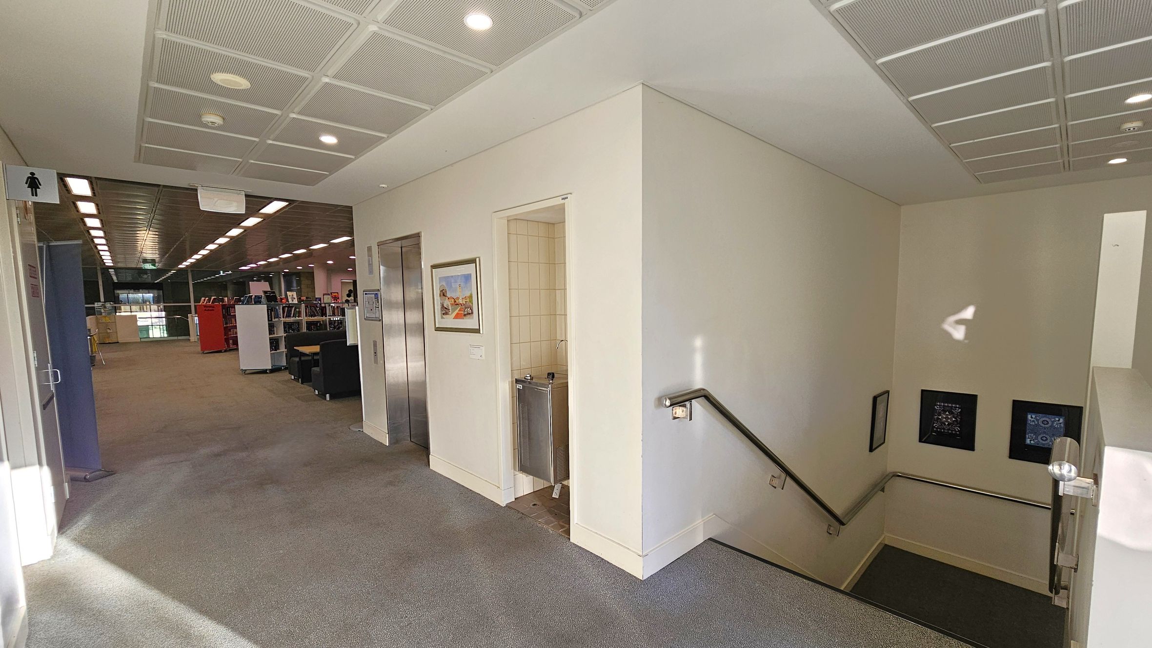 A hallway with a carpeted floor and a tiled ceiling. On the left, there's a sign indicating a women's restroom. Further down, an open area with bookshelves and seating is visible. In the centre, an elevator door is next to a water fountain and a framed picture. On the right, a staircase with a metal handrail leads downwards, with two small framed items on the wall. The area is well-lit with ceiling lights.
