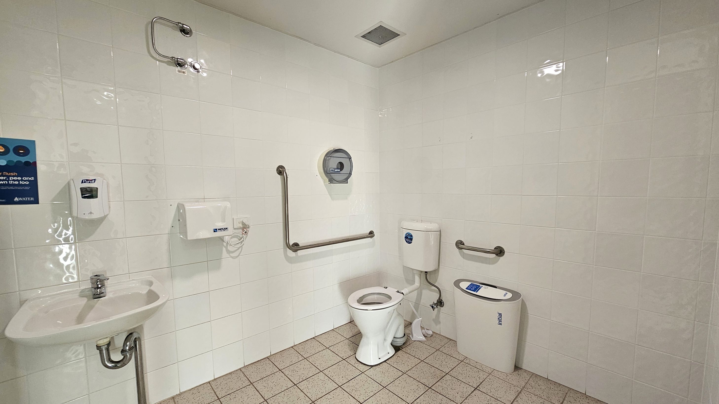 A public restroom with light-coloured tiled walls and a tiled floor. On the left, there is a small sink with a metal tap, a soap dispenser above it, and a hand dryer nearby. A poster is visible on the wall. In the centre, a toilet with a white seat is positioned against the wall, with a toilet paper dispenser above it. Metal grab bars are installed around the toilet. On the right, a sanitary disposal bin is placed next to the toilet. The ceiling has a vent.